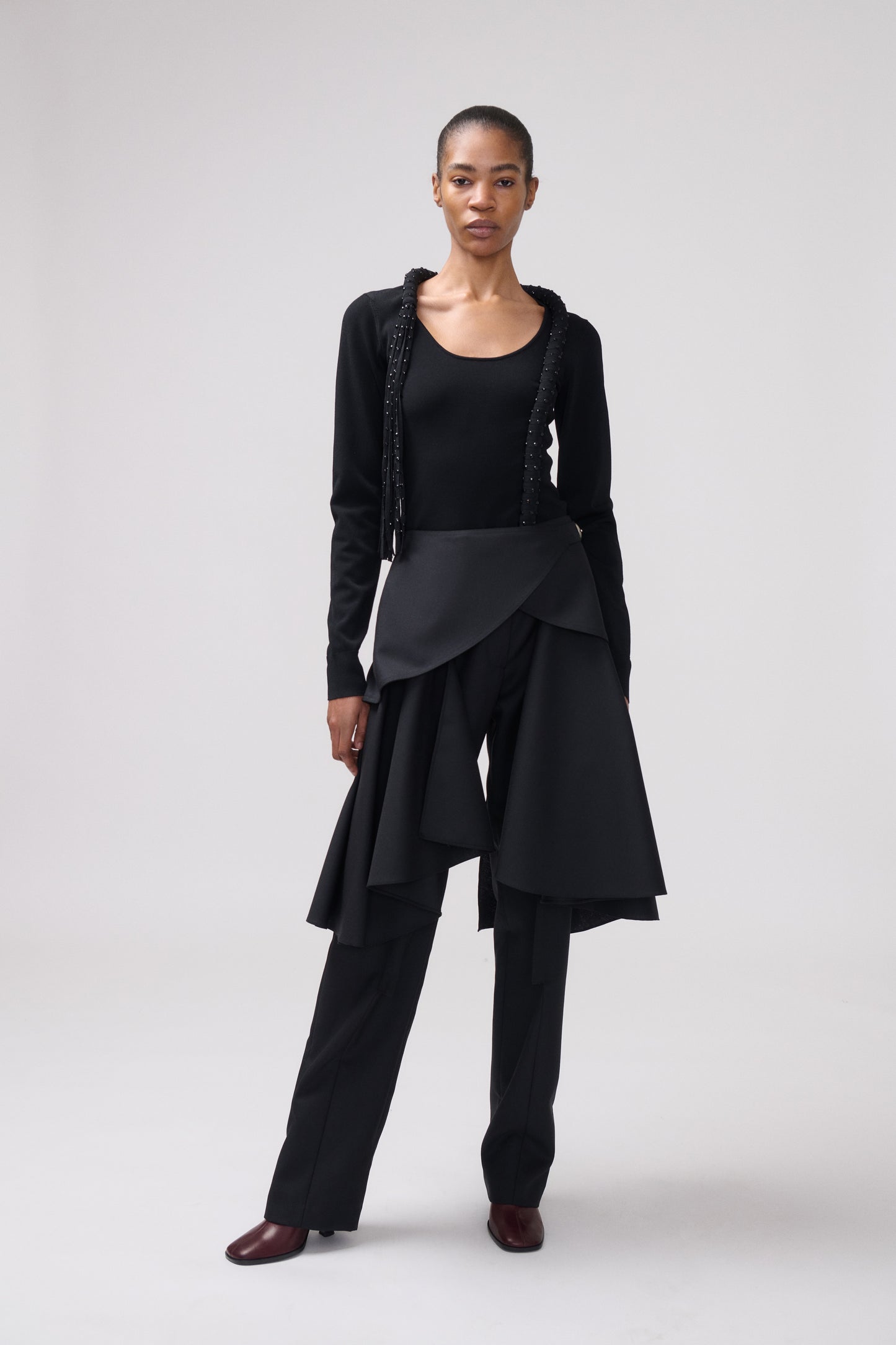 A model stands against a light gray studio background wearing a black outfit consisting of a long-sleeve top with decorative trim, a draped wrap-style skirt, matching wide-leg pants, and dark brown pointed shoes.