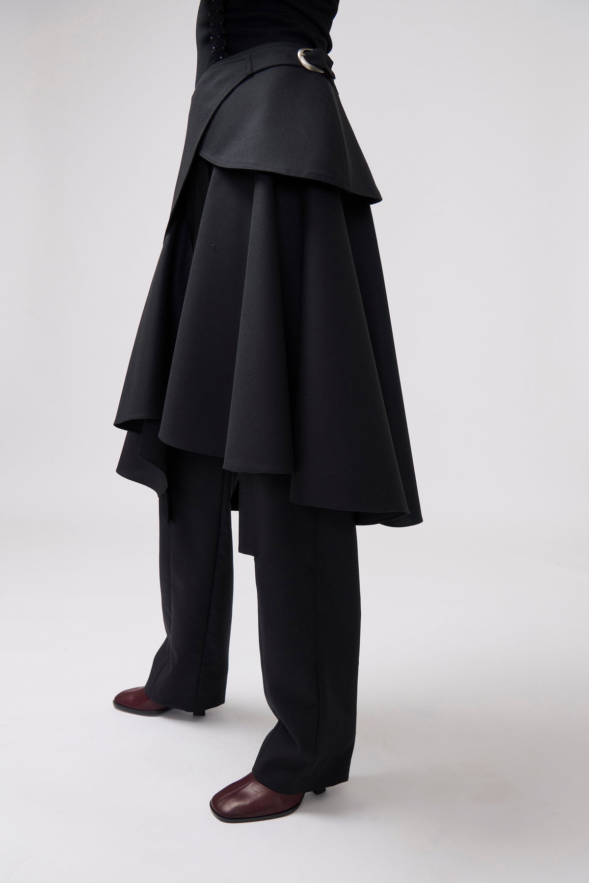 A model is shown from the waist down wearing a dramatic black asymmetrical layered skirt with a matching belt, black wide-leg trousers, and dark brown leather shoes against a light gray studio background.