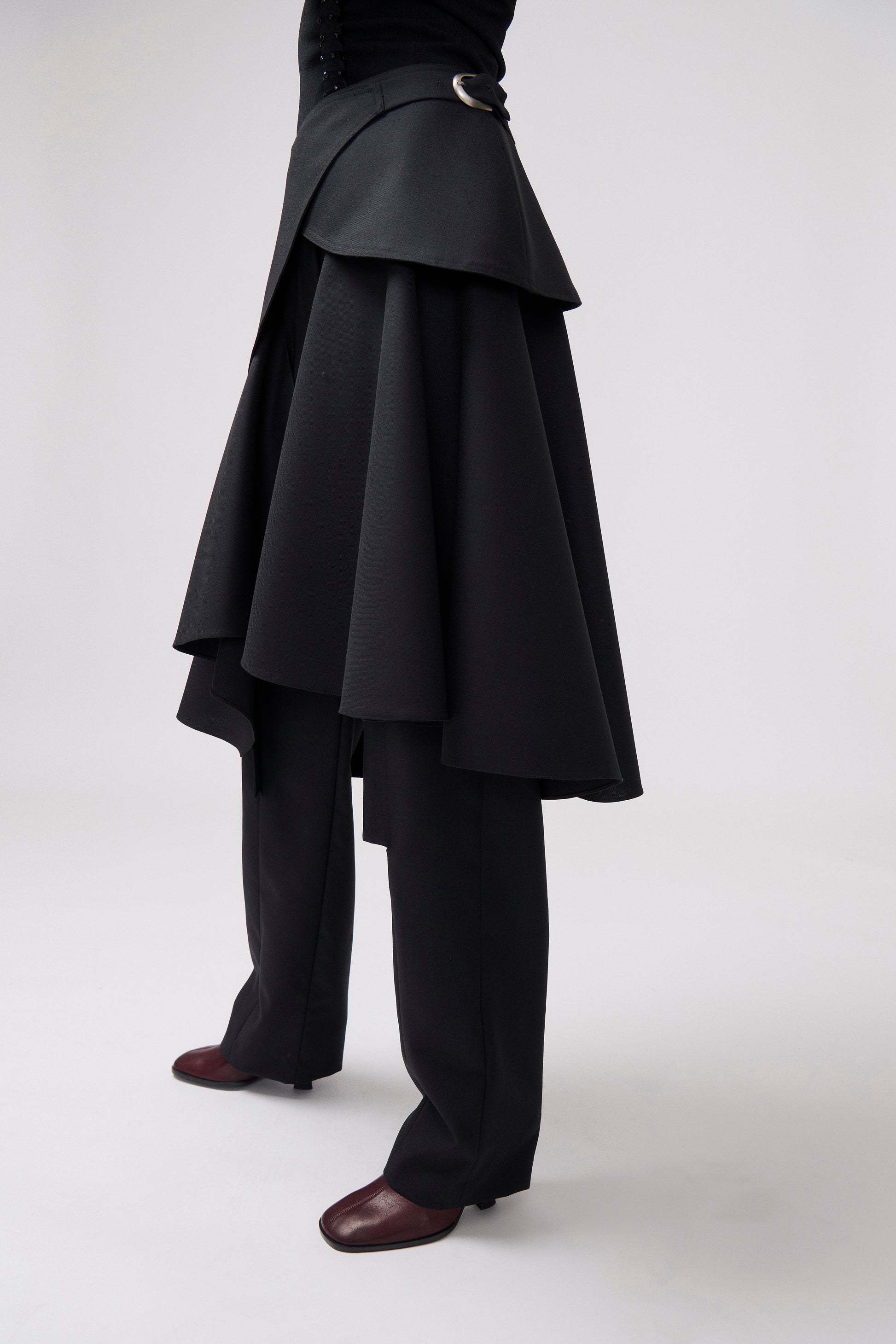 A model is shown from the waist down wearing a dramatic black asymmetrical layered skirt with a matching belt, black wide-leg trousers, and dark brown leather shoes against a light gray studio background.