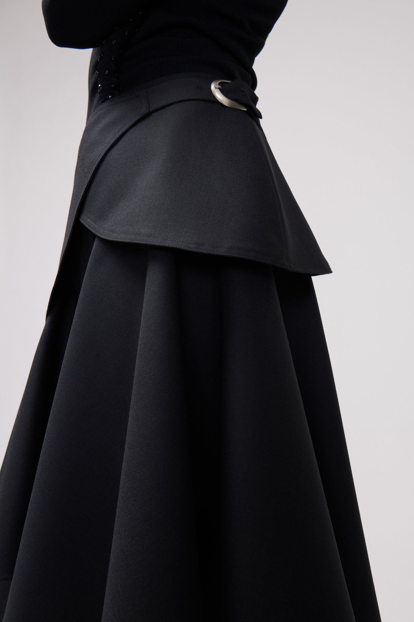 Close-up of a black trousers with a structured layered overlay and a silver-tone ring detail at the waist, shown against a light gray studio background.