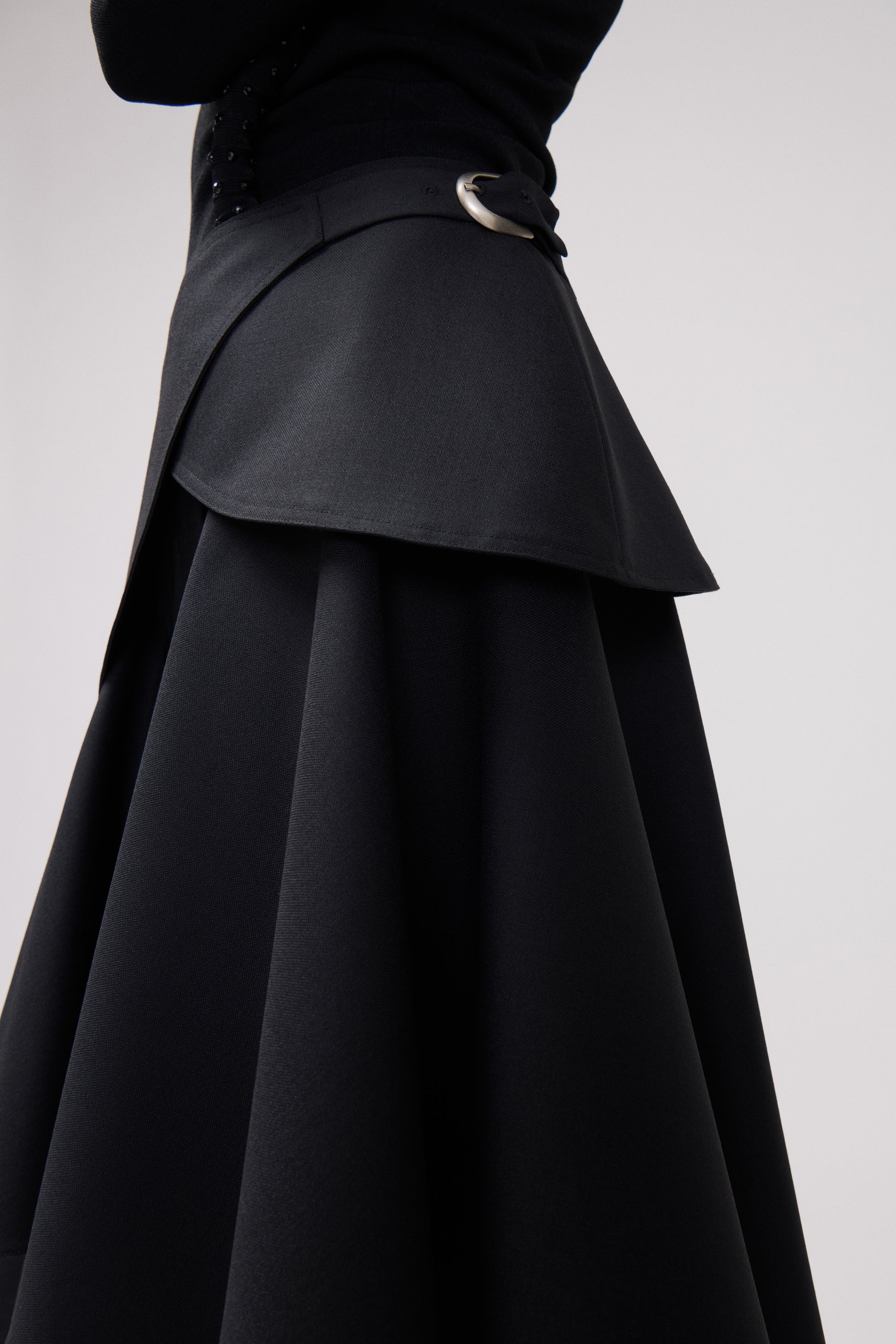 Close-up of a black trousers with a structured layered overlay and a silver-tone ring detail at the waist, shown against a light gray studio background.