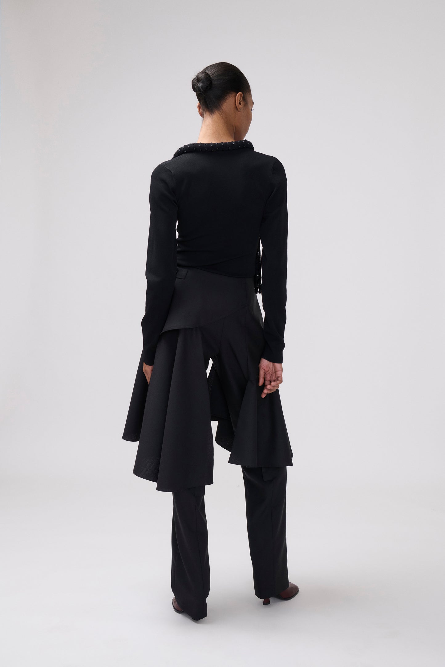 A model shown from the back wearing a black long-sleeve top with a ruffled collar, a black asymmetrical draped skirt, and black trousers, set against a plain light gray studio background.