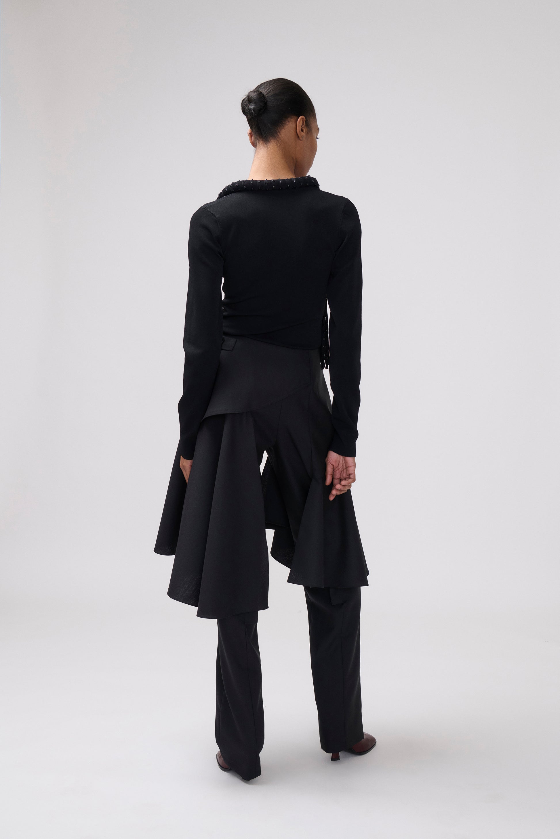 A model shown from the back wearing a black long-sleeve top with a ruffled collar, a black asymmetrical draped skirt, and black trousers, set against a plain light gray studio background.