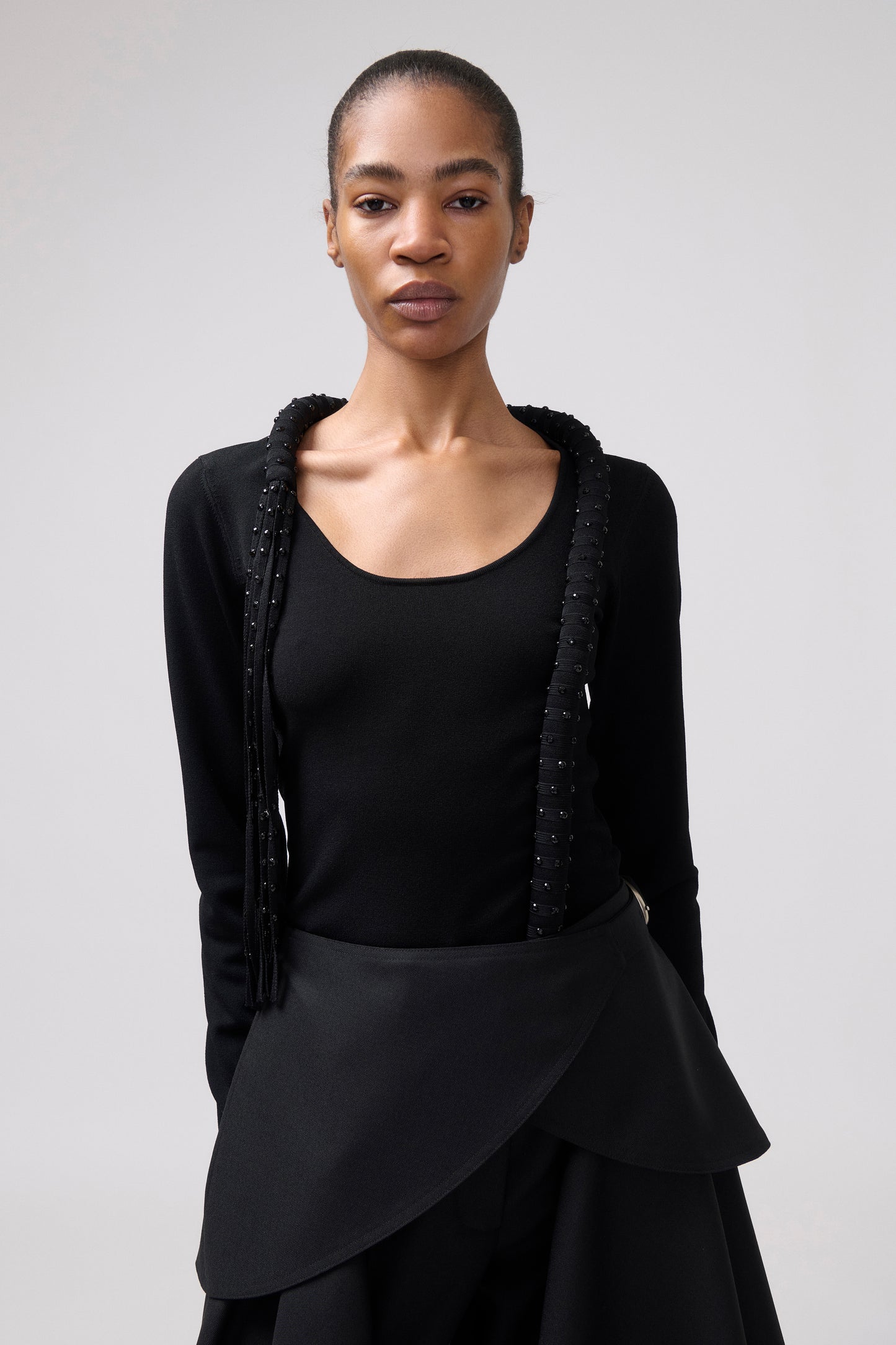 A model wearing a black long-sleeve top with textured, beaded fringe details along the shoulders and a matching black asymmetrical skirt, set against a plain light gray studio background.