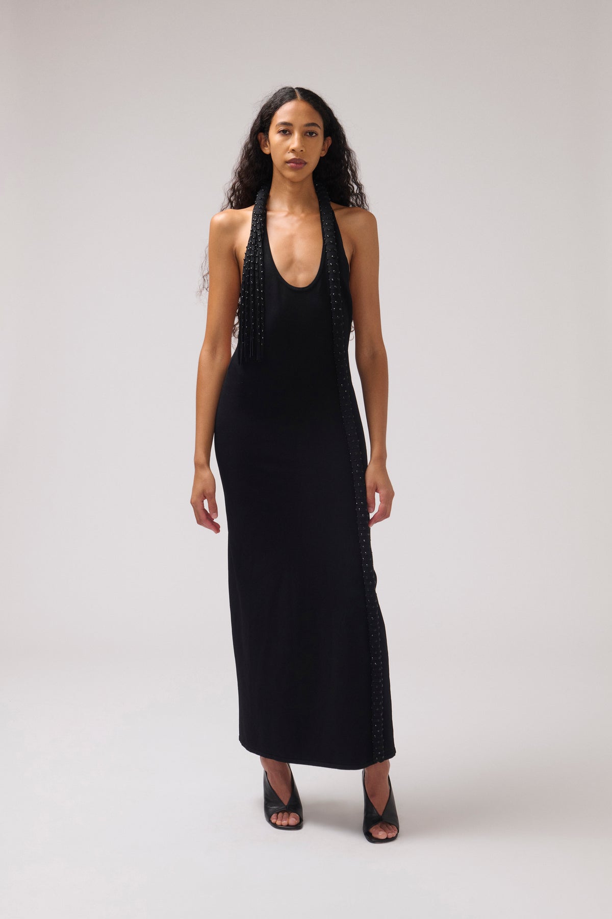 A woman modeling a black halter-neck maxi dress with beaded fringe detailing along the neckline and side seams, paired with black strappy heels, against a neutral light gray studio background.