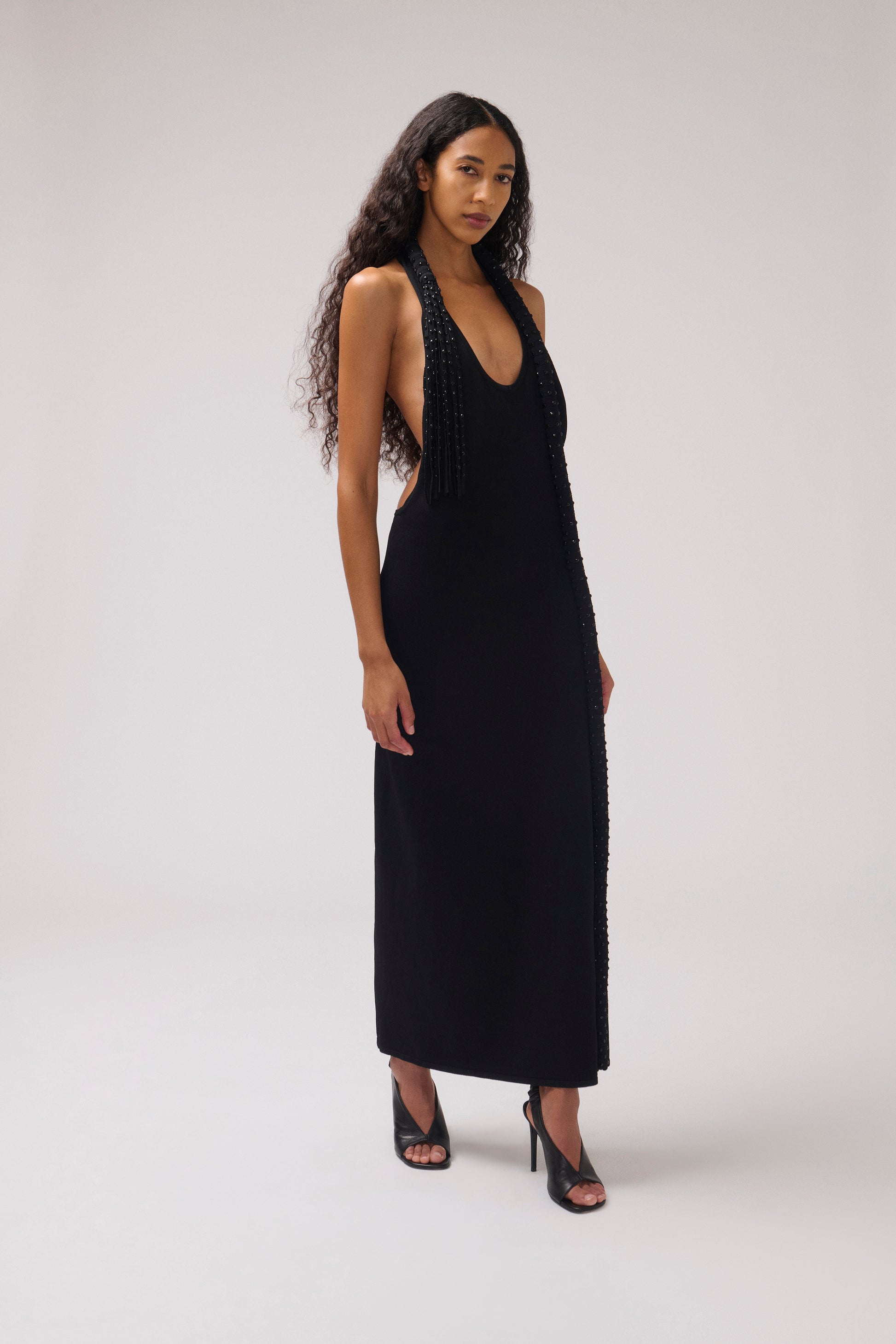 A woman modeling a sleeveless black maxi dress with beaded fringe detailing along the neckline and side seams, paired with black high-heeled sandals, against a light gray studio background.