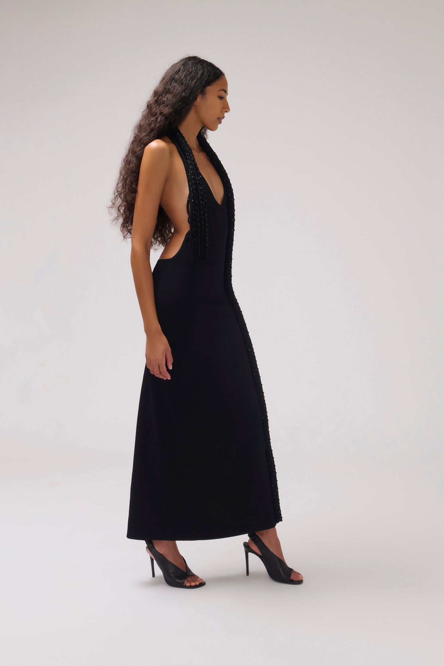 A woman modeling a sleeveless black maxi dress with a deep V-neckline, beaded halter-style neckline, and side cutouts, paired with black high-heeled sandals against a light gray studio background.