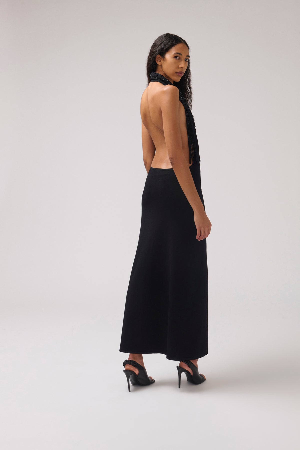 A woman modeling a black halter-neck maxi dress with an open back, shown from the side against a light gray studio background.
