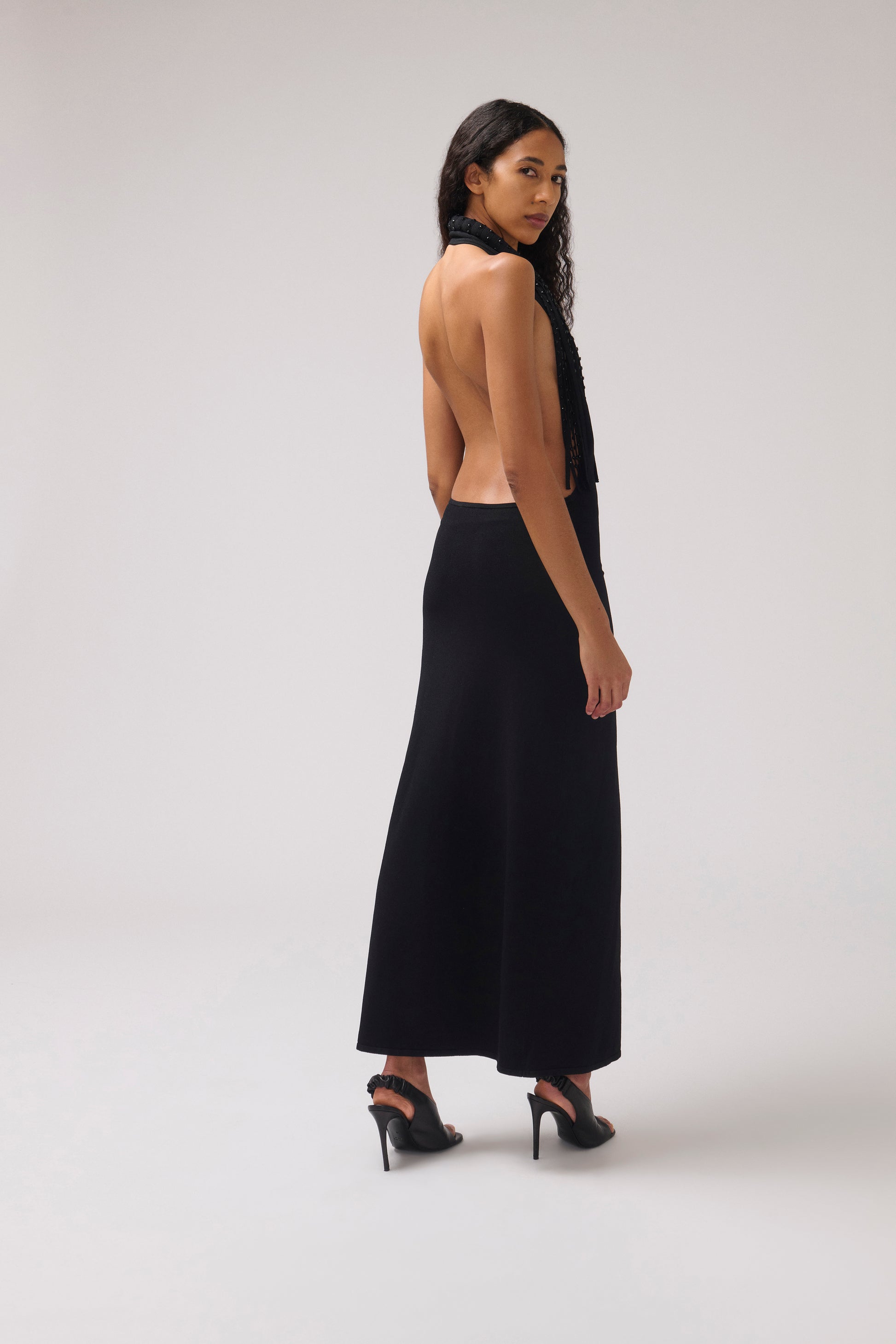 A woman modeling a black halter-neck maxi dress with an open back, shown from the side against a light gray studio background.