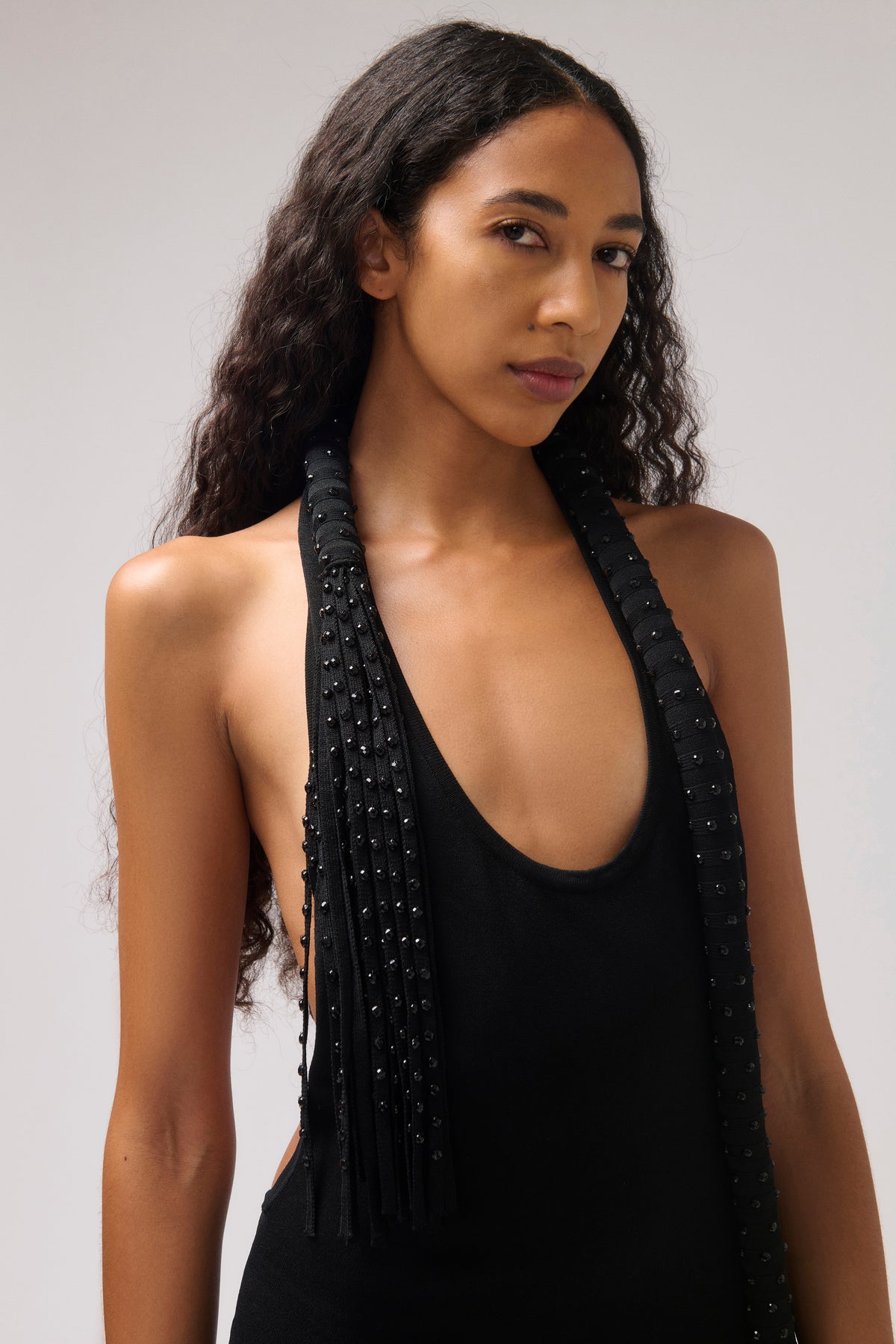 A woman modeling a black halter-neck top with beaded fringe details along the straps, photographed against a neutral light gray studio background.