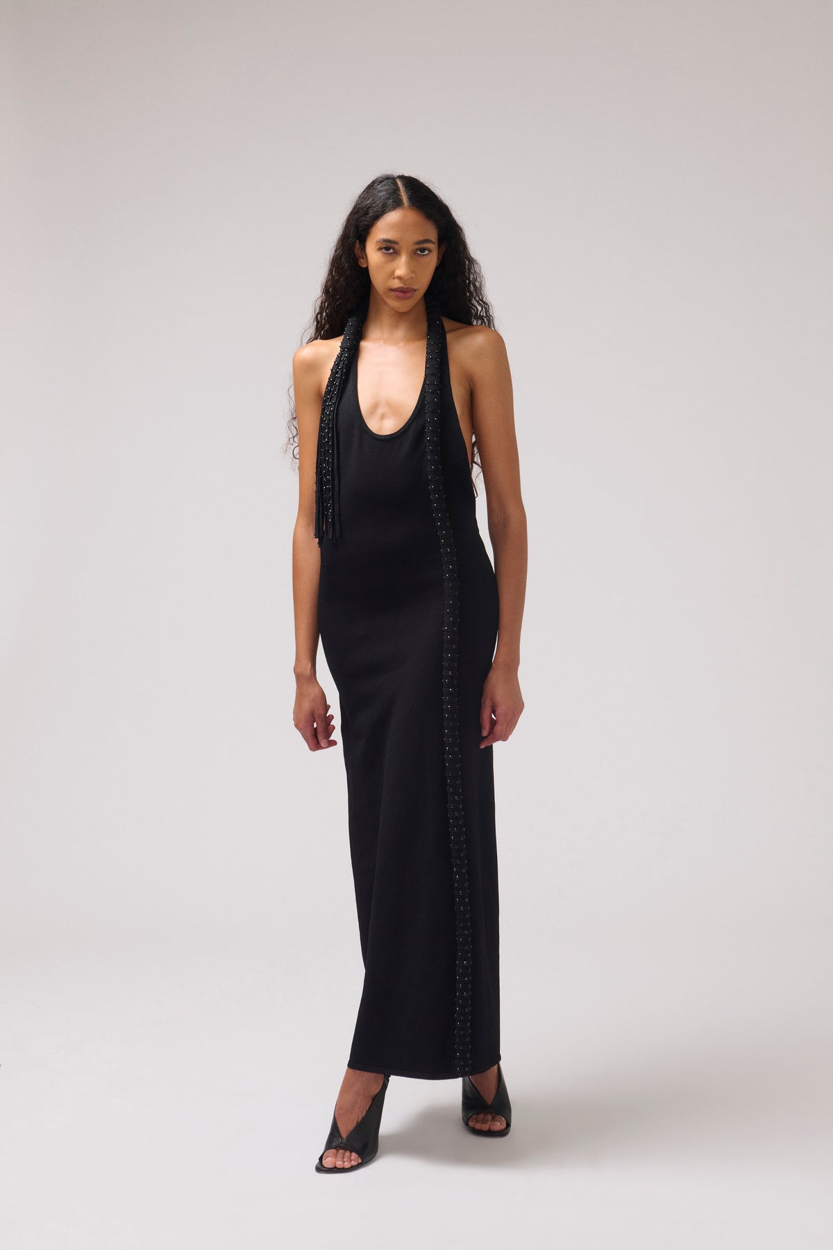 A model wearing a black sleeveless maxi dress with decorative beadwork down the front and sides, paired with black strappy heels, against a light gray studio background.