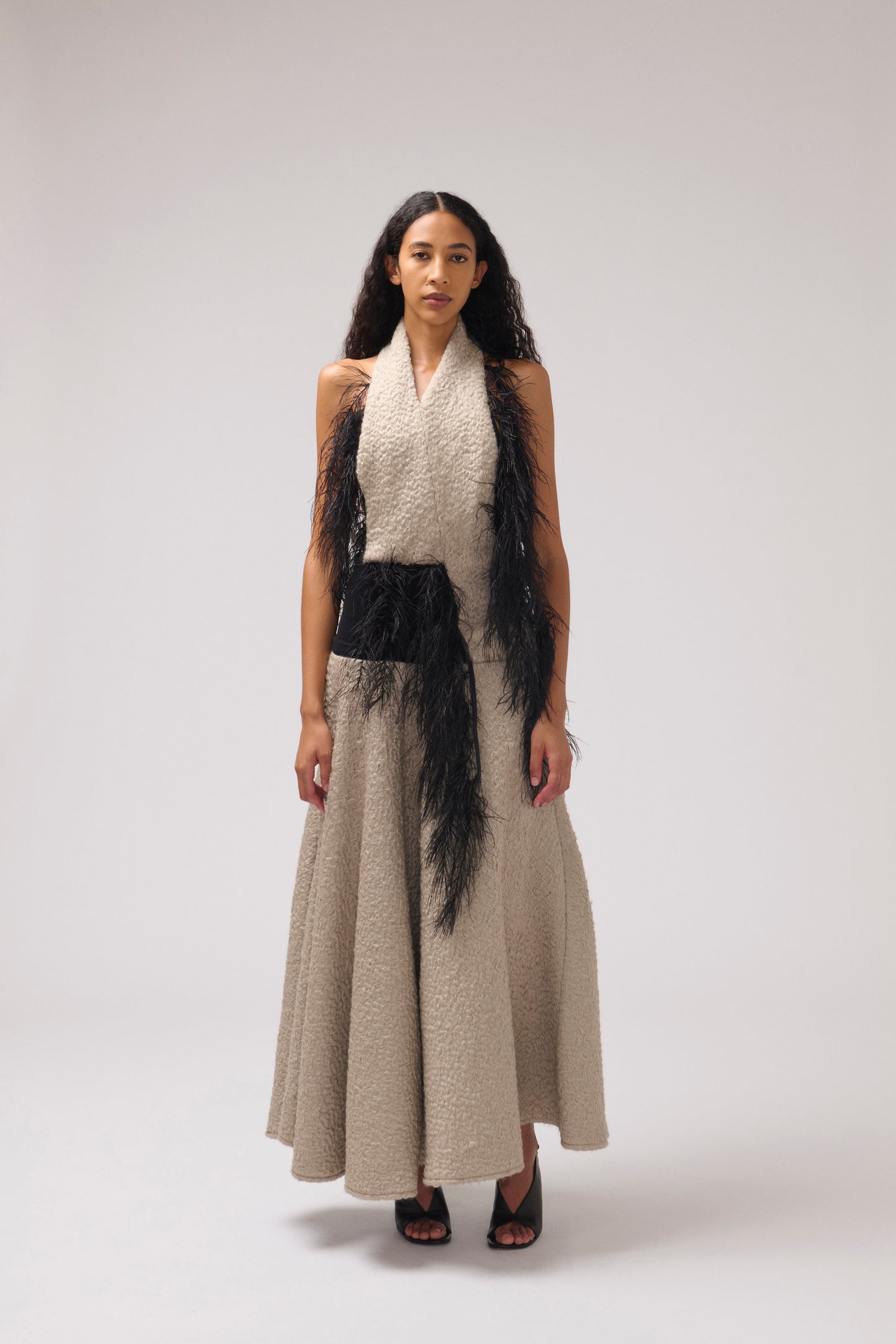 A model stands against a light gray studio background wearing a halterneck, textured parchment maxi dress with a wide collar, a black feathered detail, and black strappy heels.