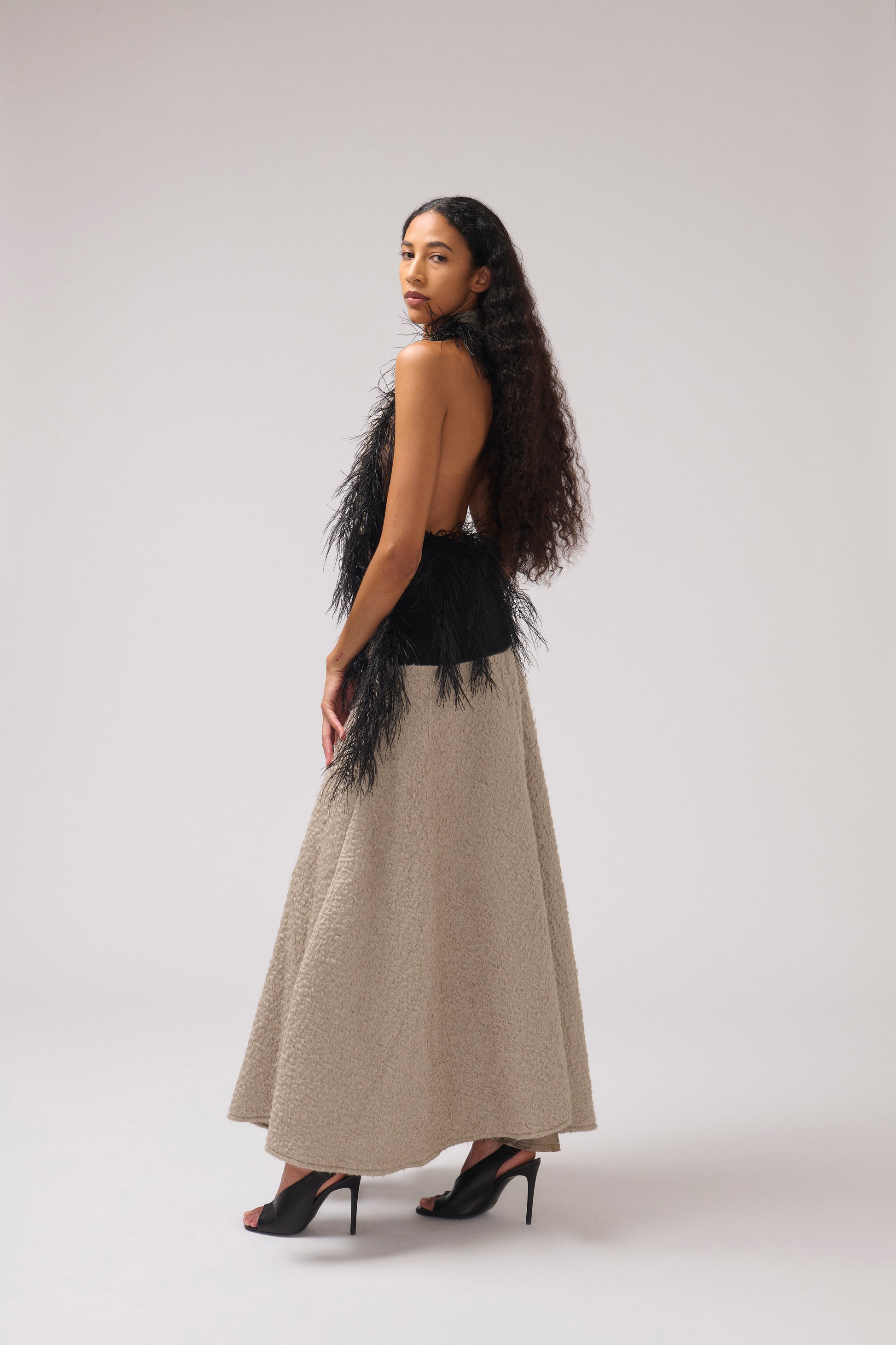 A model standing sideways against a light gray studio background wearing a halterneck, textured parchment maxi dress with a wide collar, a black feathered detail, and black strappy heels.