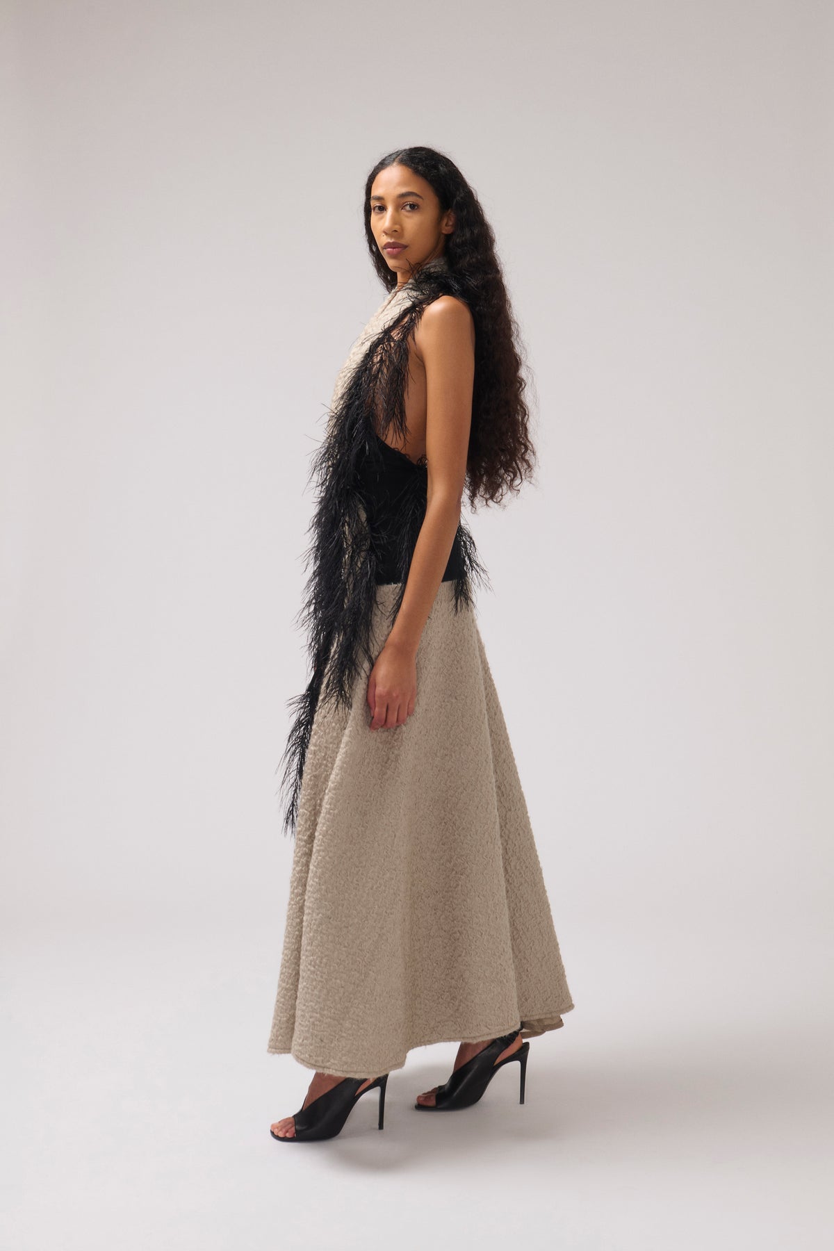 A model is shown from the side in a studio setting, wearing a beige halter dress with black feather trim paired with black high heels.