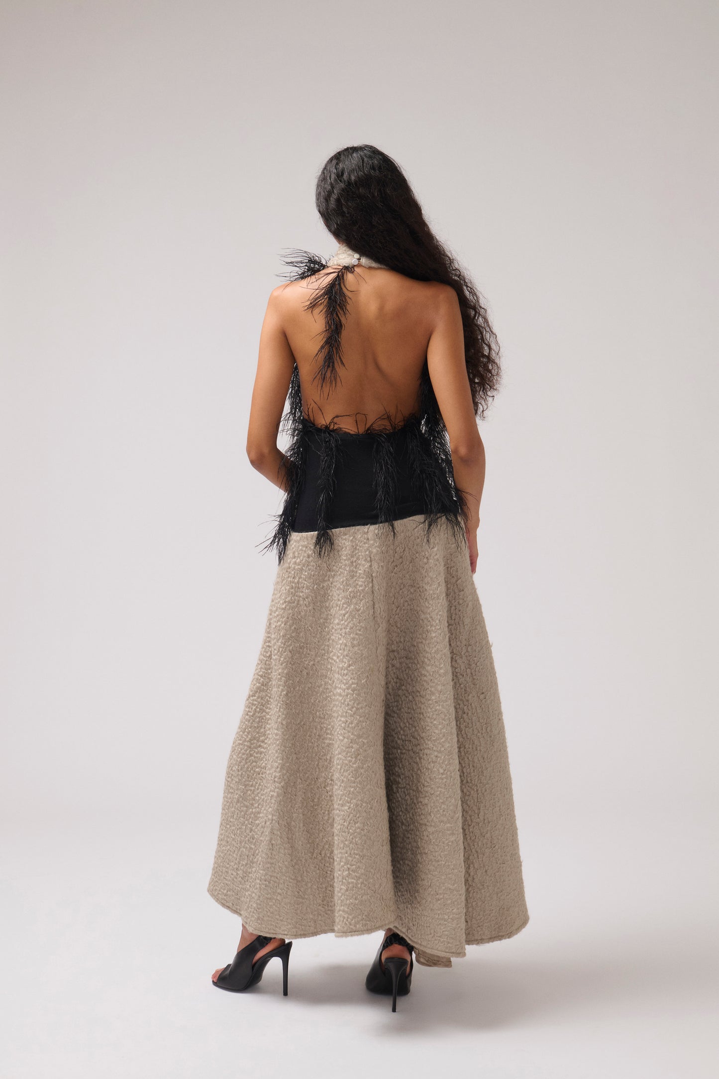 A model pictured from the back, stands against a light gray studio background wearing a halterneck, textured parchment maxi dress with a wide collar, a black feathered detail, and black strappy heels.