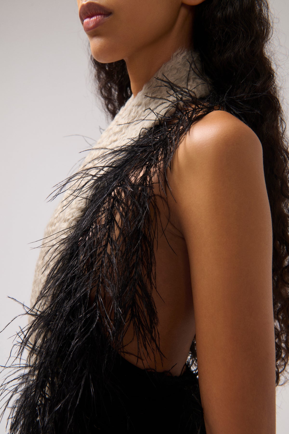 Close-up of a model wearing a beige halter dress with dramatic black feather trim, shown against a light gray studio background.