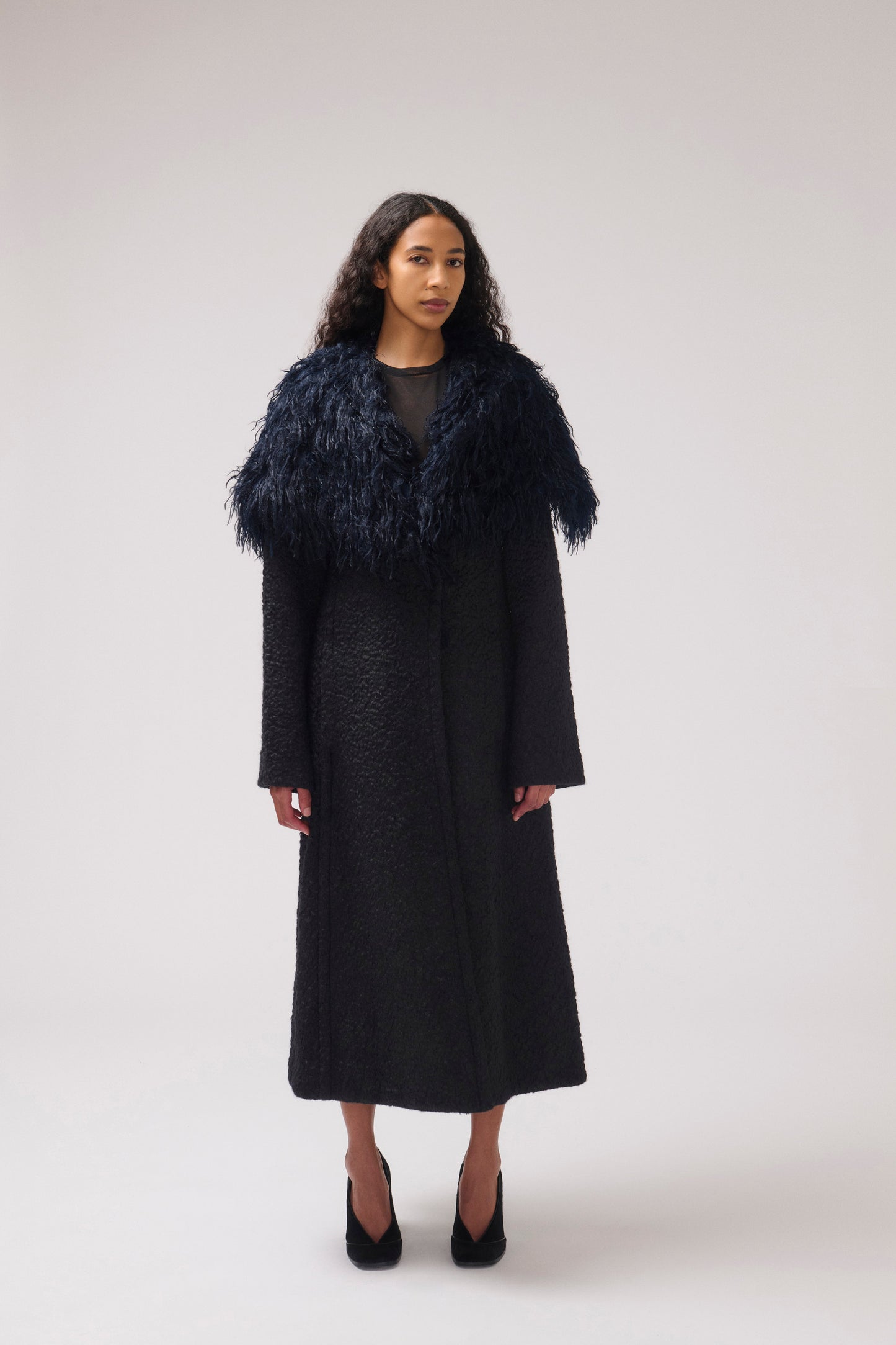 A model stands against a light gray studio background wearing a long black textured coat with a large dark navy feathered collar and black pointed heels.