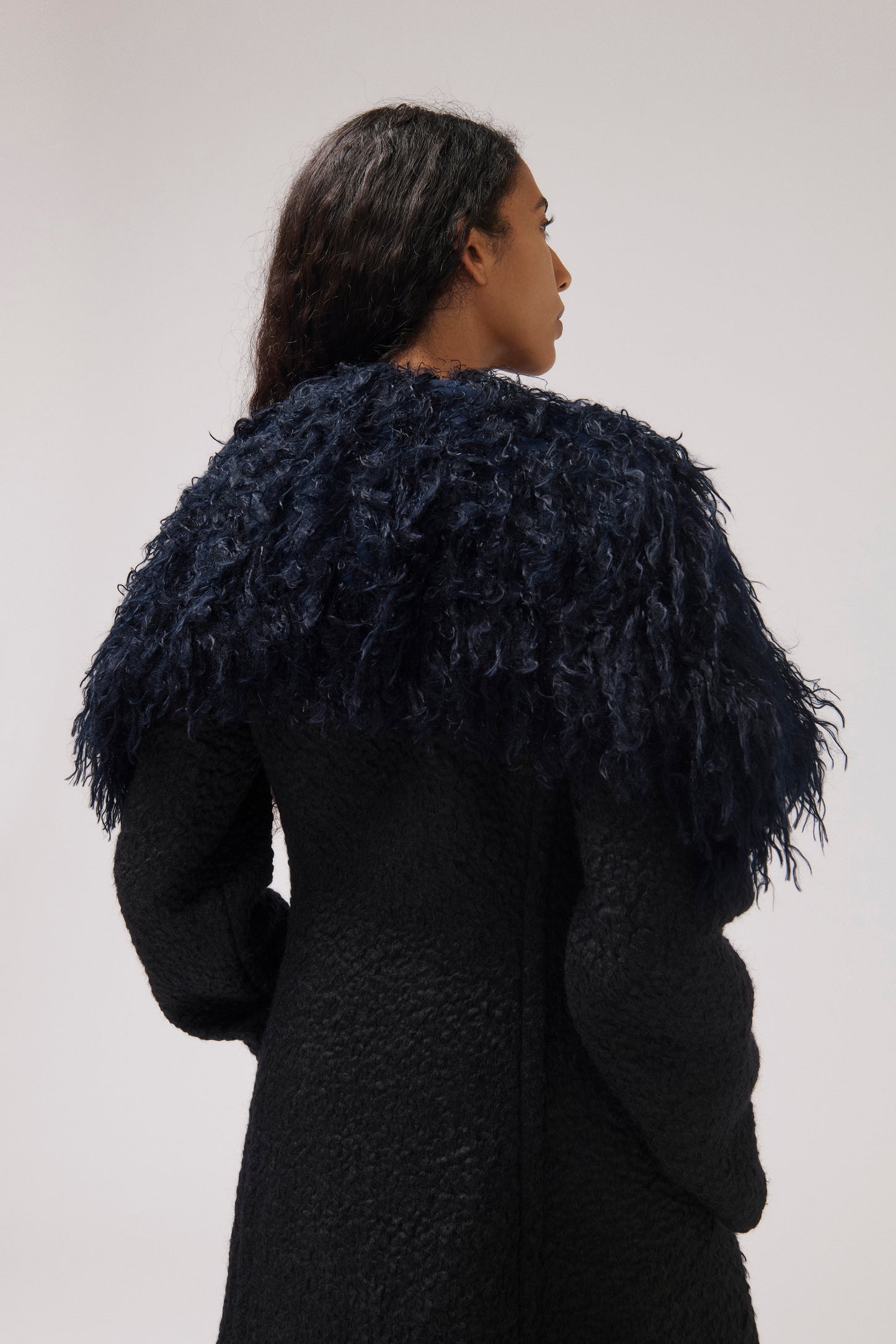 A woman shown from the back wearing a dark textured coat with a voluminous dark navy feathered collar against a plain light gray studio background.