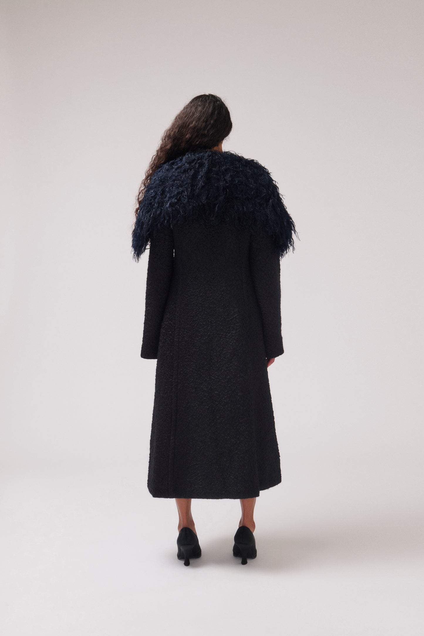 A model viewed from behind wearing a long black textured coat with a dramatic dark navy feathered collar, paired with black heels, against a neutral light gray studio background.
