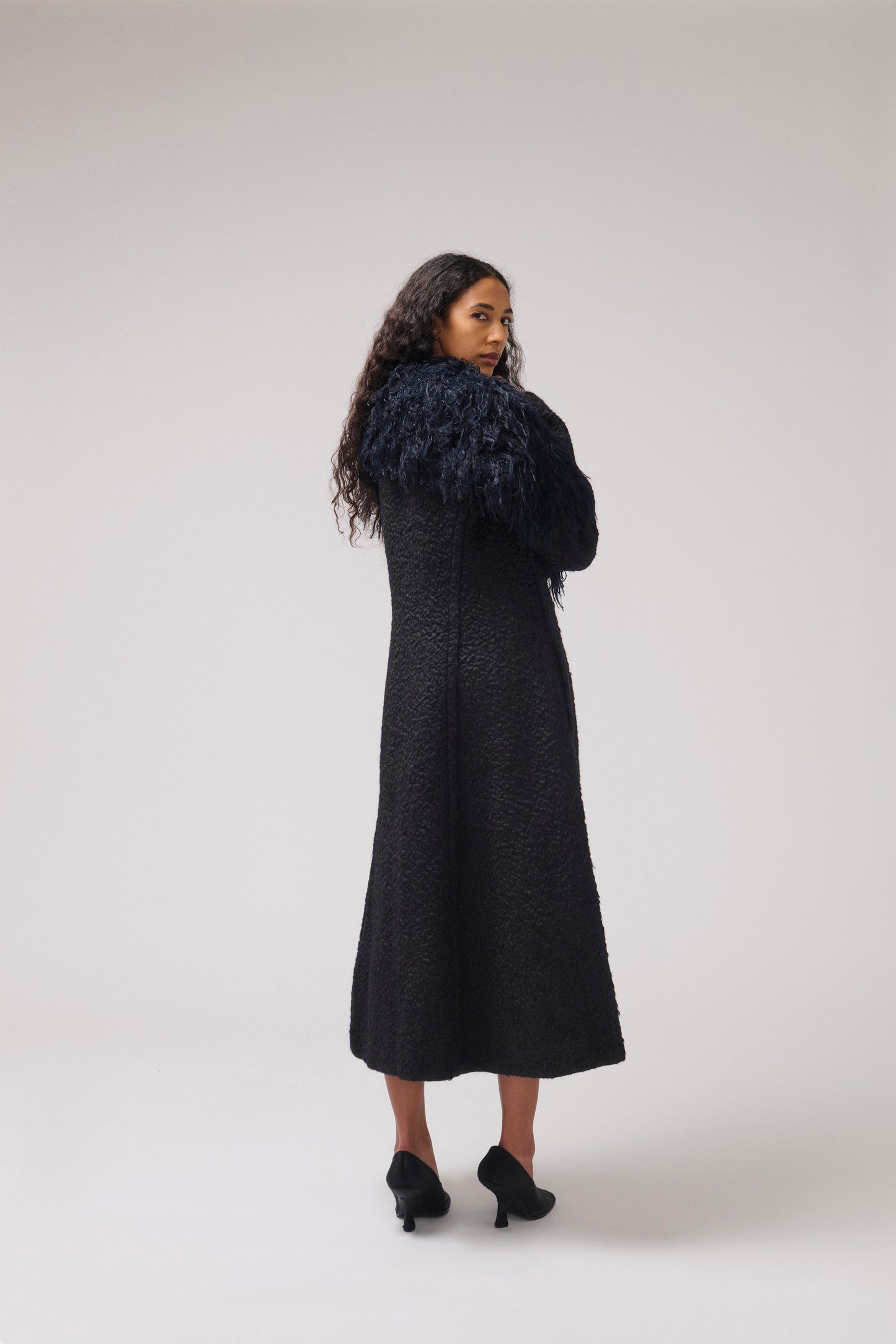 A woman stands in a studio wearing a long black textured midi dress with a voluminous dark blue feathered collar and black heels, photographed against a light gray background.