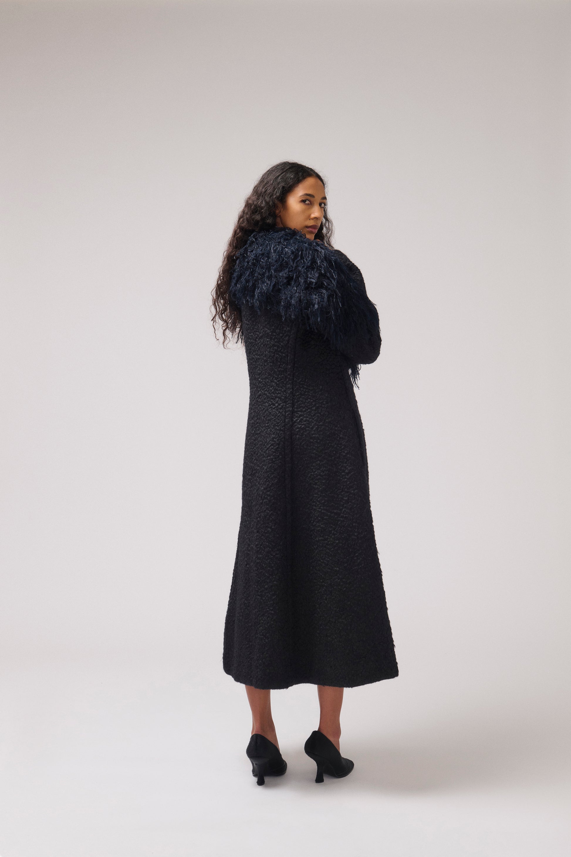 A woman stands in a studio wearing a long black textured midi dress with a voluminous dark blue feathered collar and black heels, photographed against a light gray background.