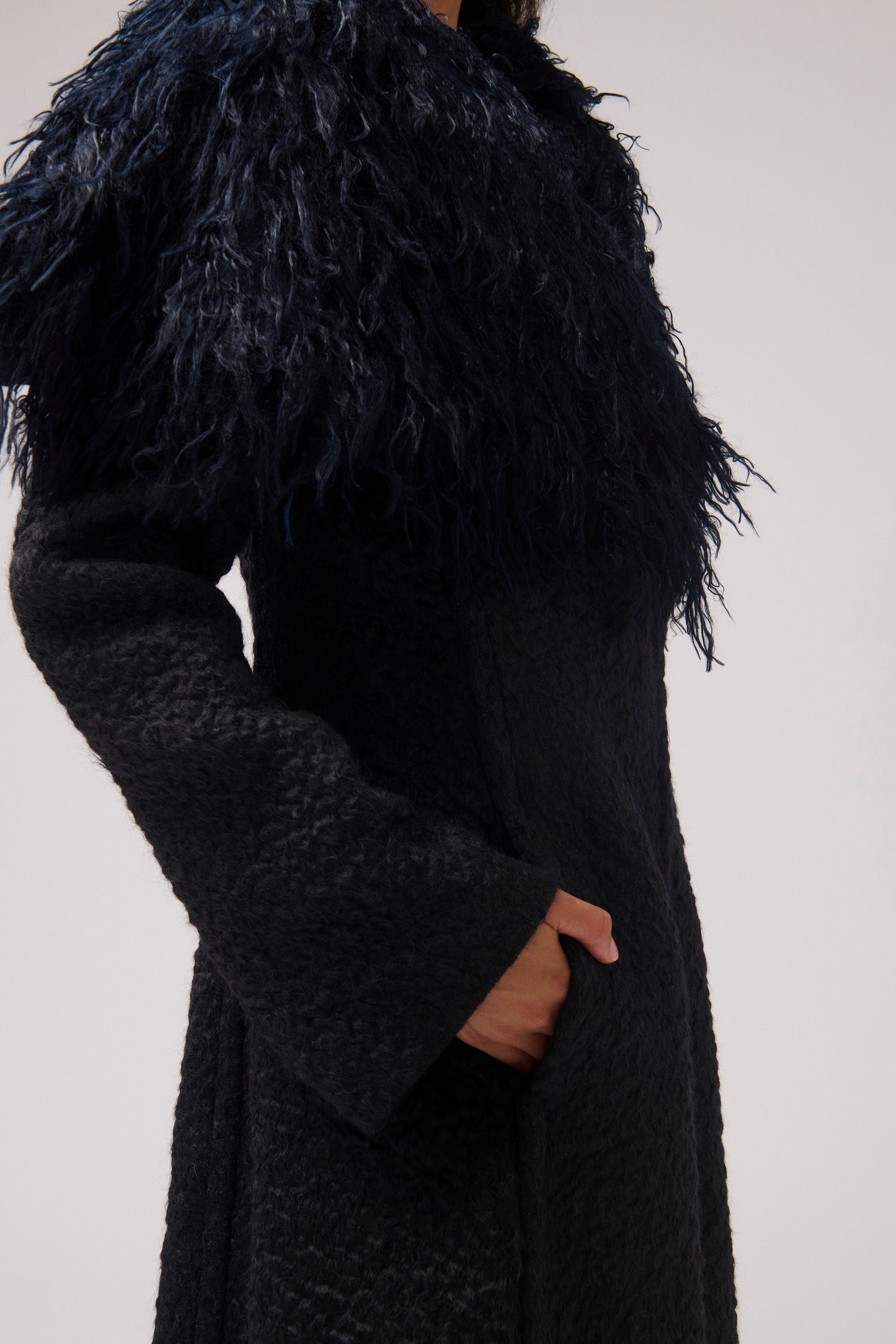 A close-up of a person wearing a long black textured coat with a dramatic dark feathered collar, shown against a plain light background.