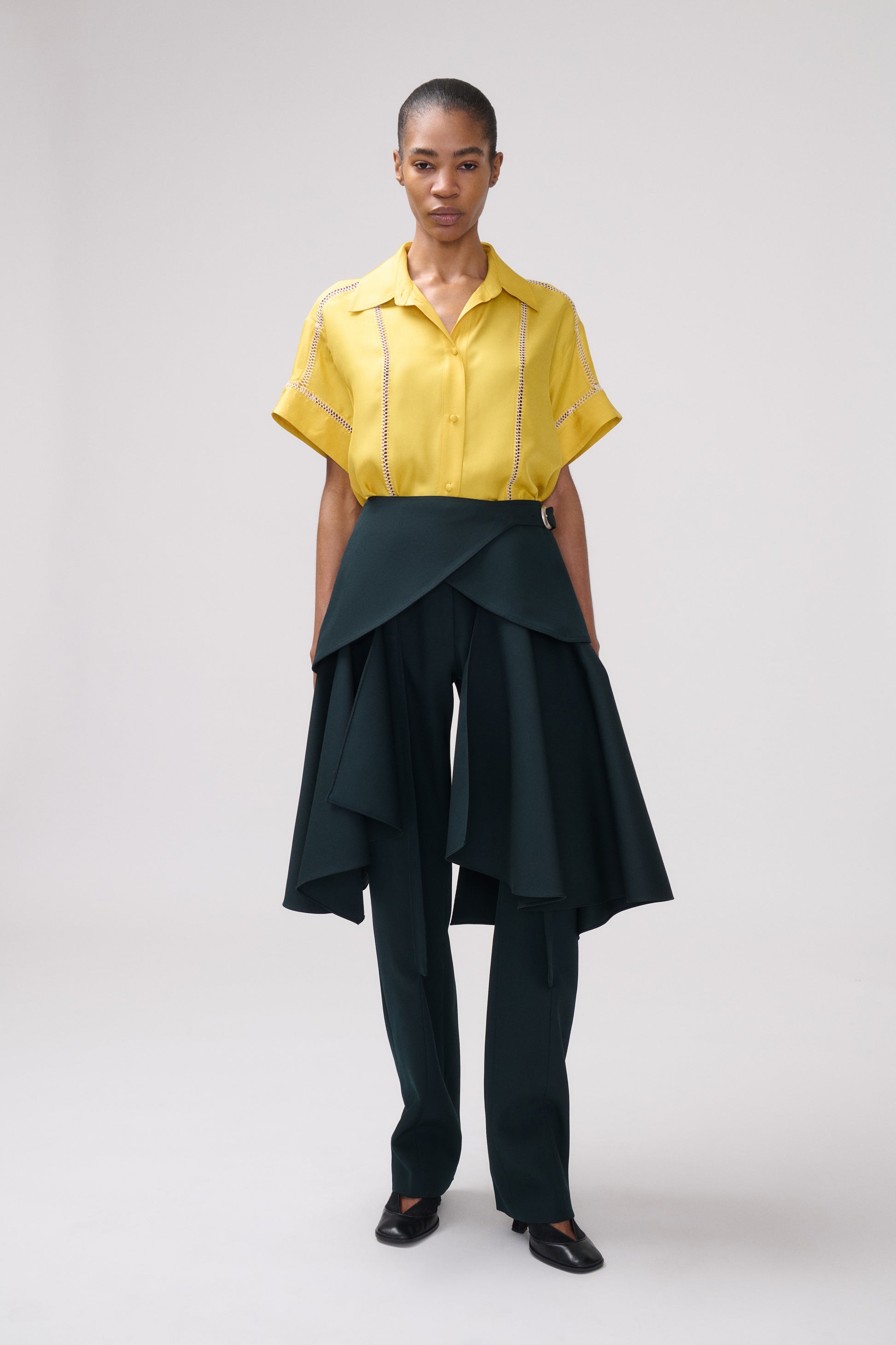 A model stands against a light gray studio background wearing a bright yellow short-sleeve button-up shirt with hand stitched detailing 