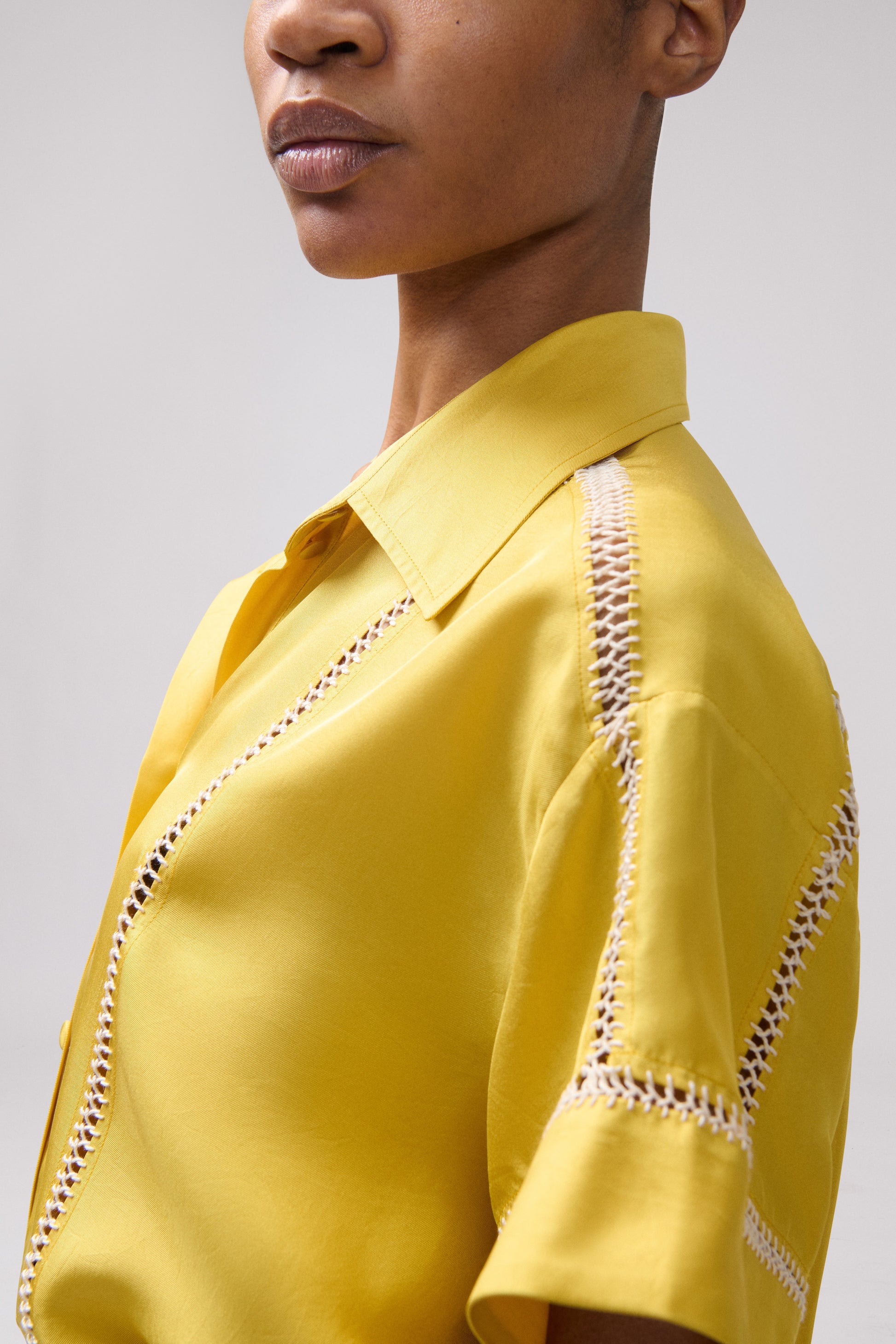 Close-up of a model wearing a bright yellow short-sleeve collared blouse with decorative cream embroidered detailing along the shoulders and sleeve.