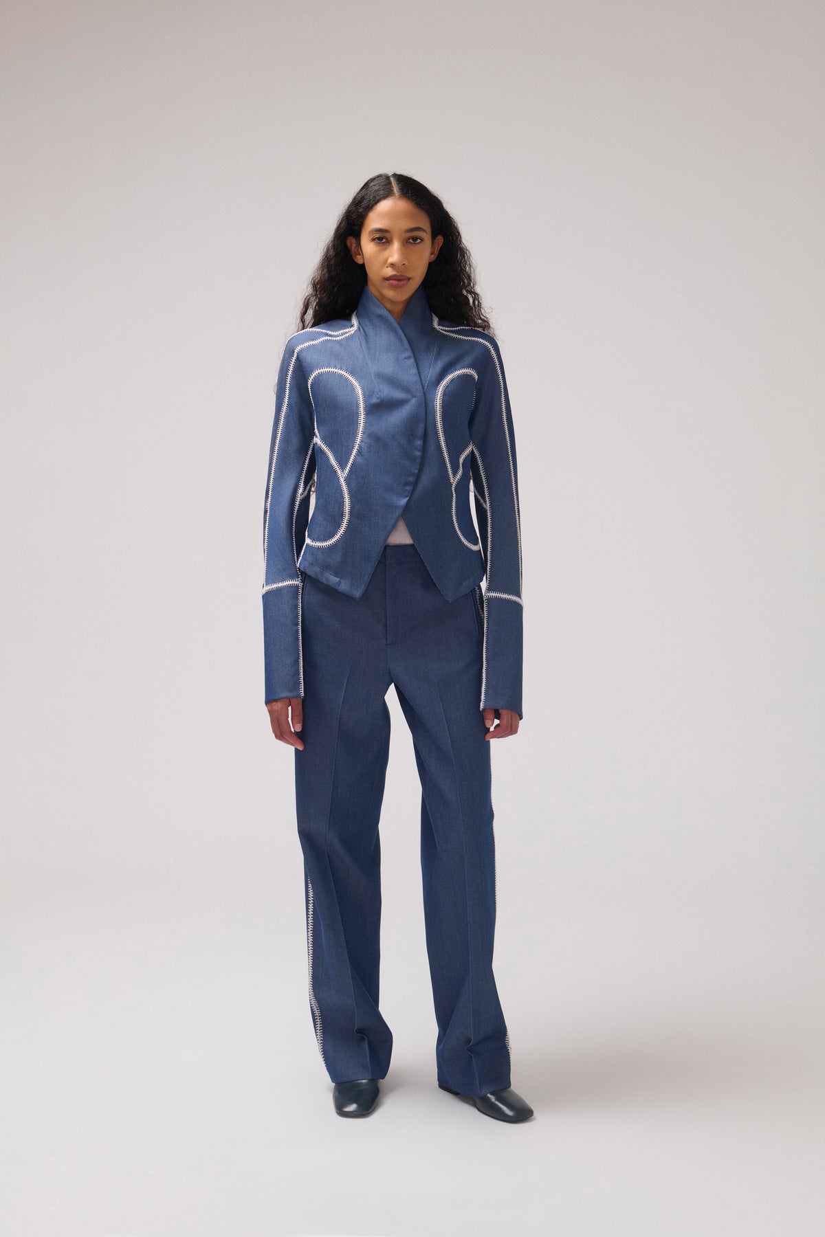 A model stands against a light gray studio background wearing a blue tailored jacket and matching wide-leg trousers with contrasting white curved piping details.