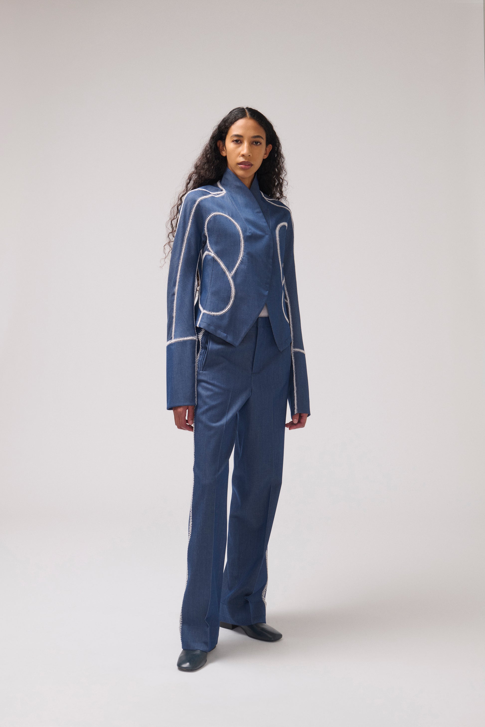 A model stands against a light gray studio background wearing a blue tailored suit with white embroidered geometric line details on the jacket and matching trousers.