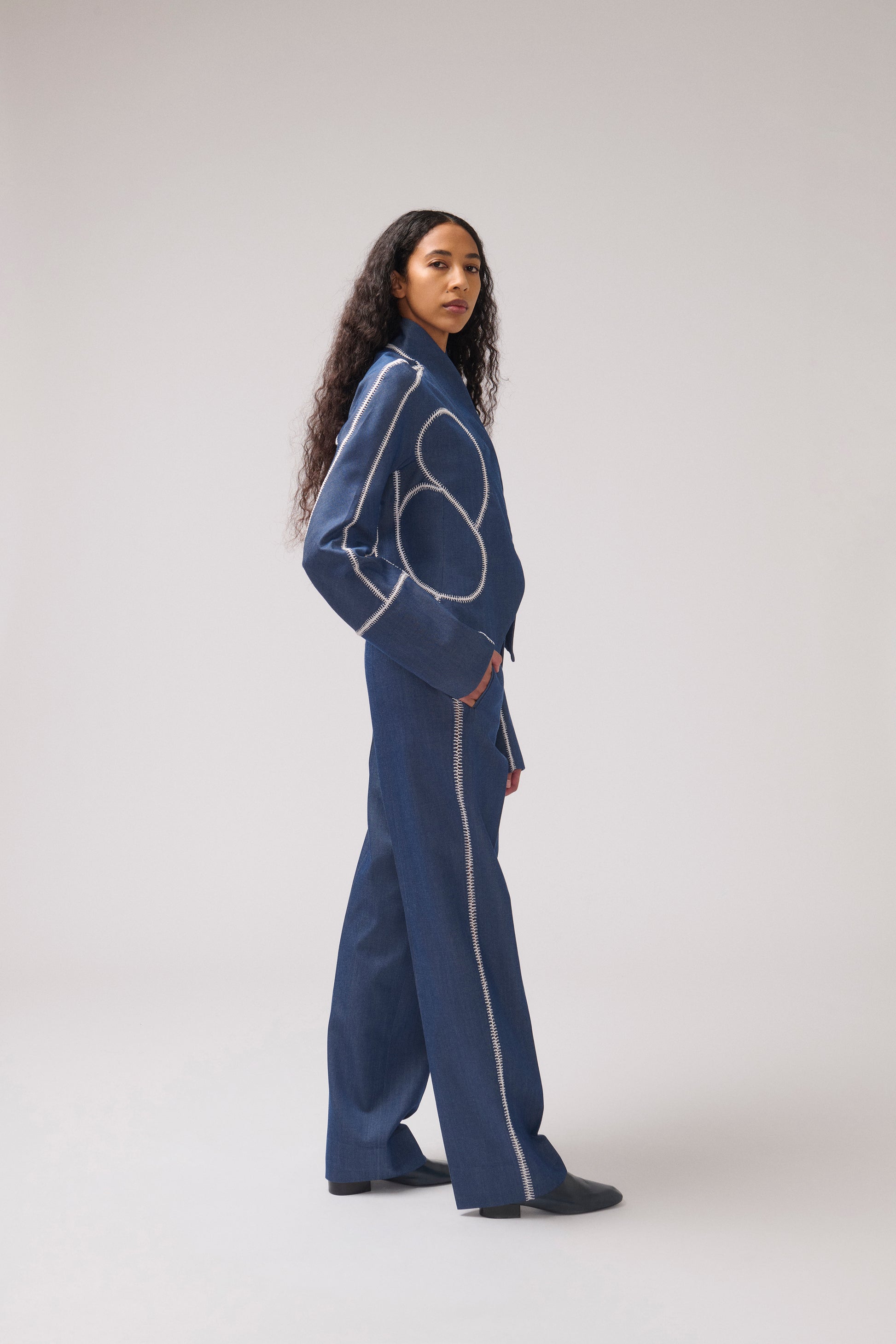 A model stands in profile wearing a dark blue wide-leg jumpsuit with contrasting light blue geometric line embroidery on the chest and along the side seams, set against a plain light gray studio background.