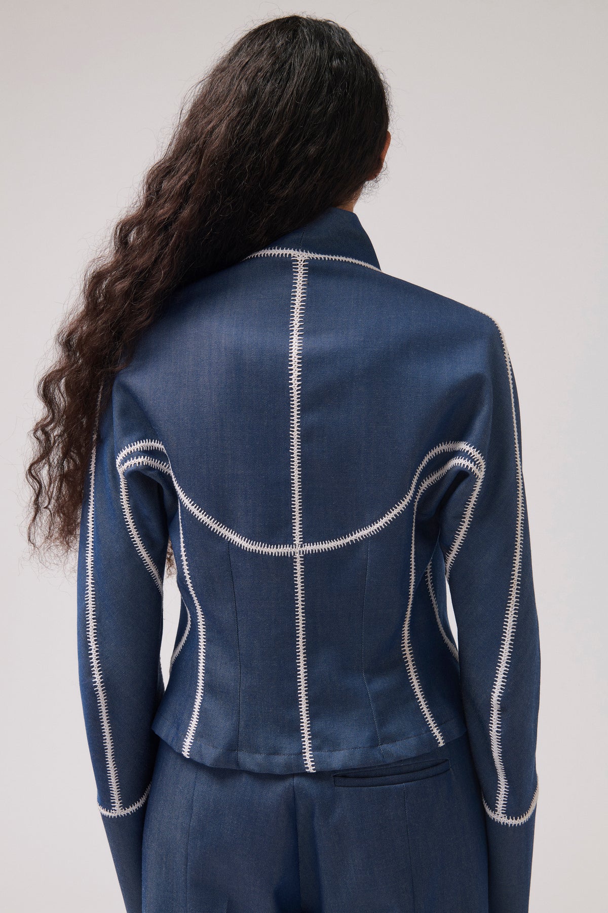 A model shown from the back wearing a navy blue cropped jacket with prominent white contrast stitching, paired with matching navy trousers, against a light gray studio background.