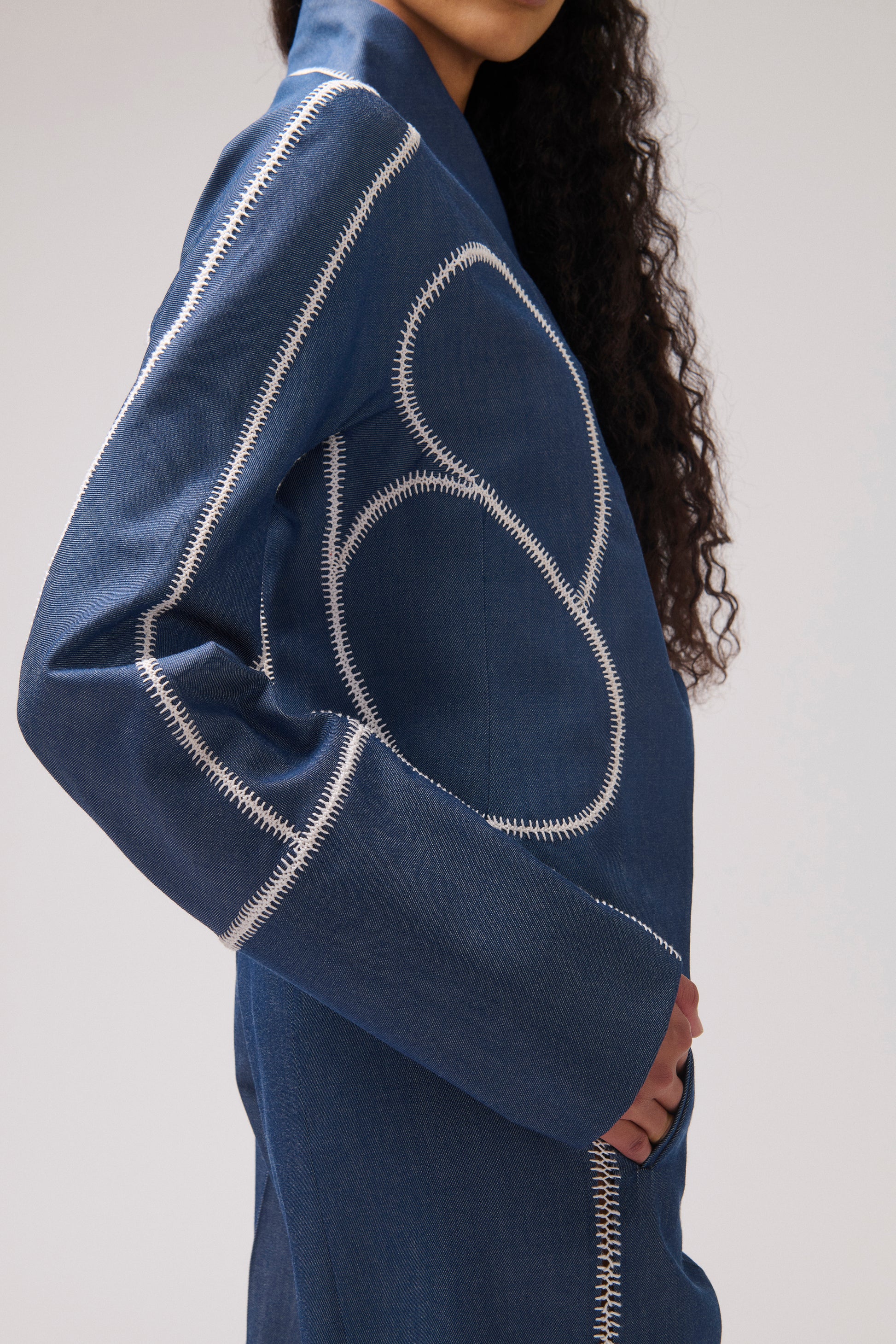 A close-up side view of a person wearing a blue denim jacket with prominent white contrast stitching and abstract curved stitching details on the sleeve and body, against a plain light gray background.