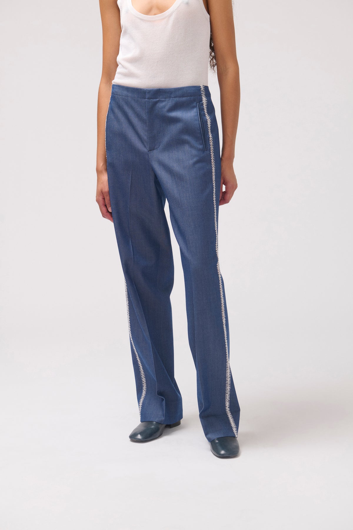 A model wearing blue wide-leg pants with white side stripes, paired with a white tank top and dark shoes, photographed against a light gray studio background.
