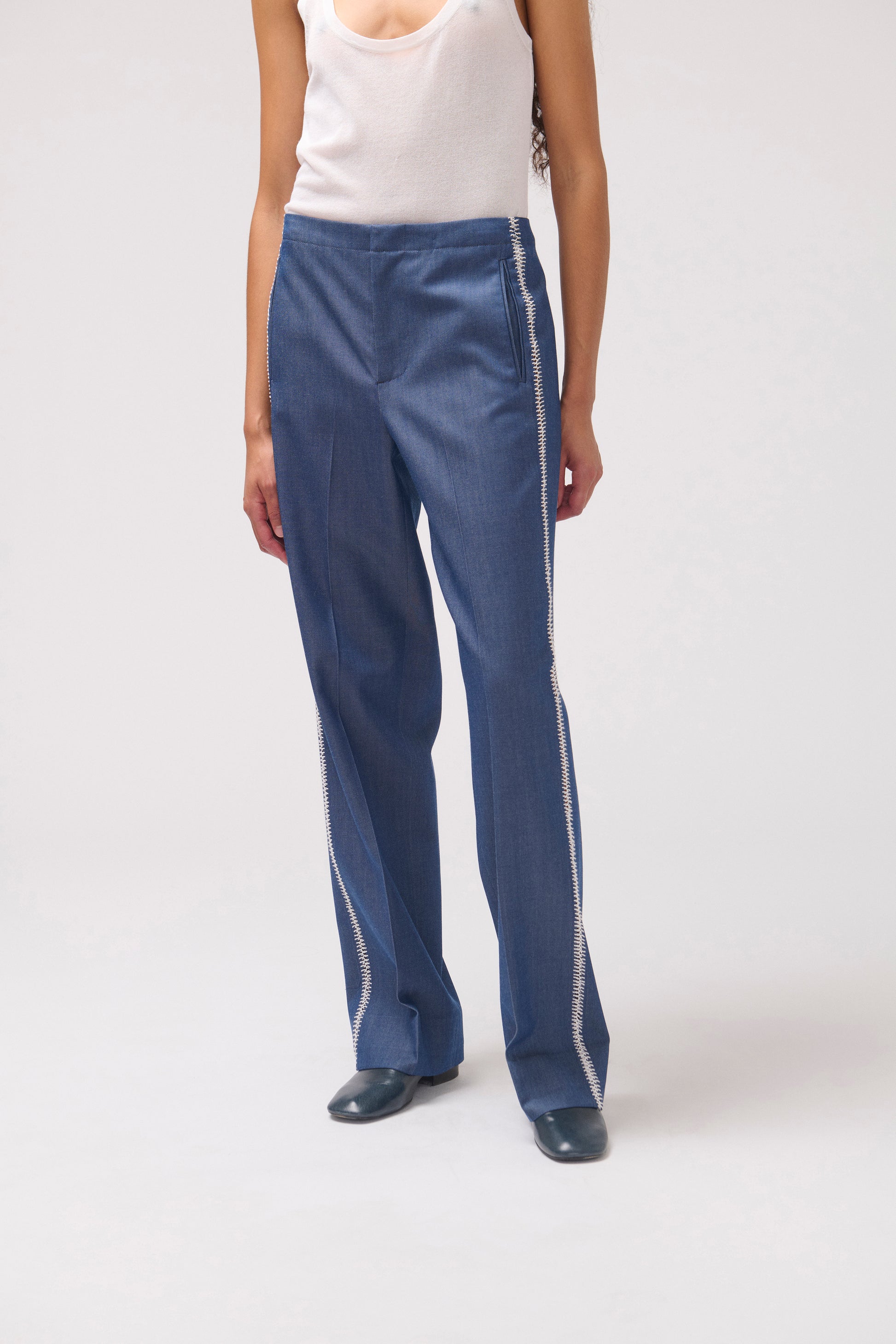 A model wearing blue wide-leg pants with white side stripes, paired with a white tank top and dark shoes, photographed against a light gray studio background.