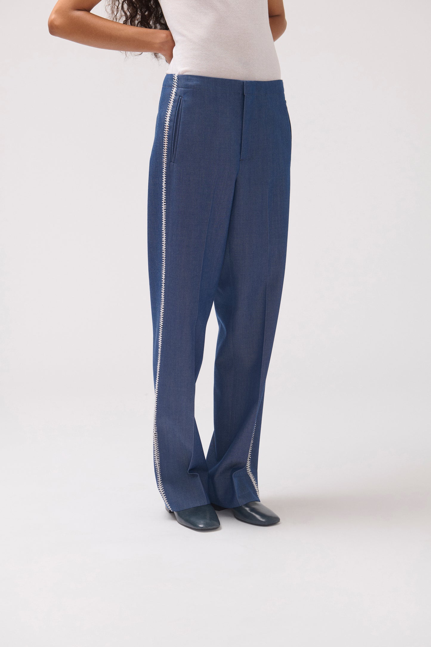 A model wearing blue wide-leg trousers with a silver chain detail running down the side, paired with a white top and dark shoes, photographed against a light gray studio background.