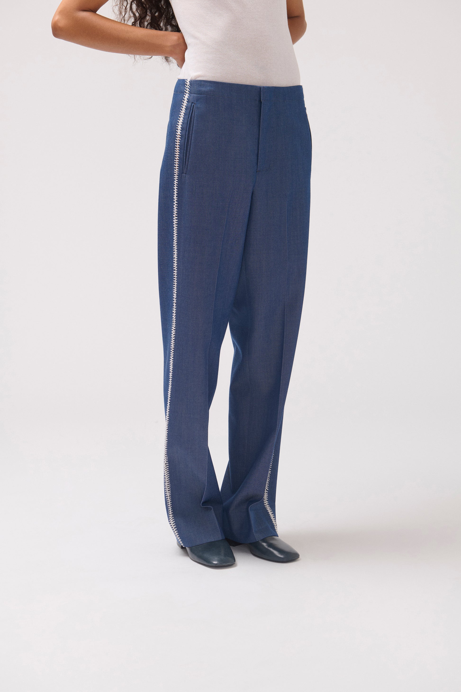 A model wearing blue wide-leg trousers with a silver chain detail running down the side, paired with a white top and dark shoes, photographed against a light gray studio background.