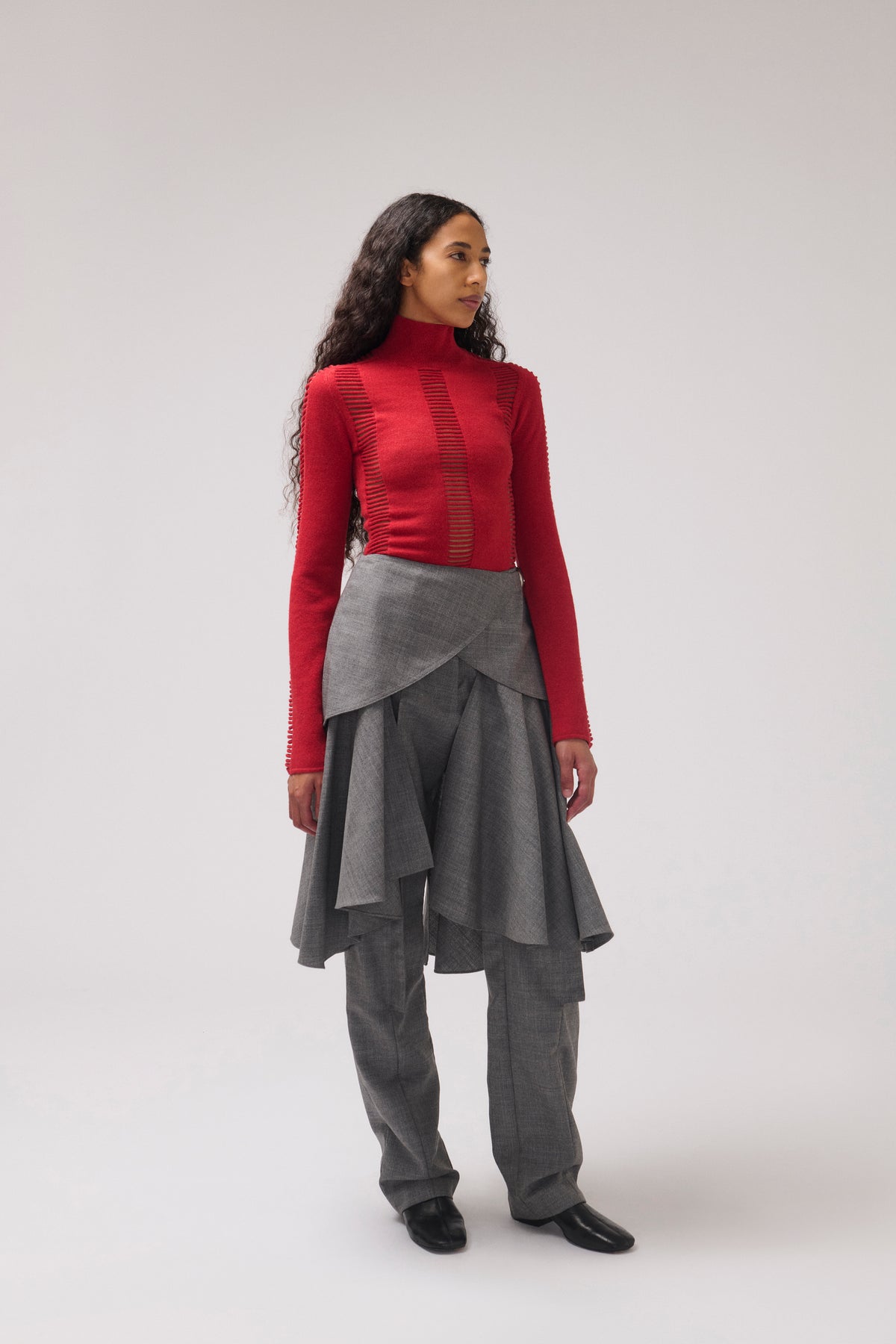 A model stands in profile wearing a bright red mock-neck long-sleeve top with vertical textured panels, paired with a layered gray wrap skirt and matching gray wide-leg trousers, against a light gray studio background.