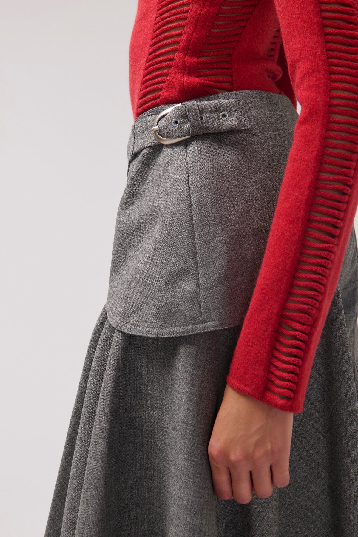 Close-up of a person wearing a gray pleated skirt with a matching belt and a red ribbed long-sleeve top, shown against a light gray studio background.