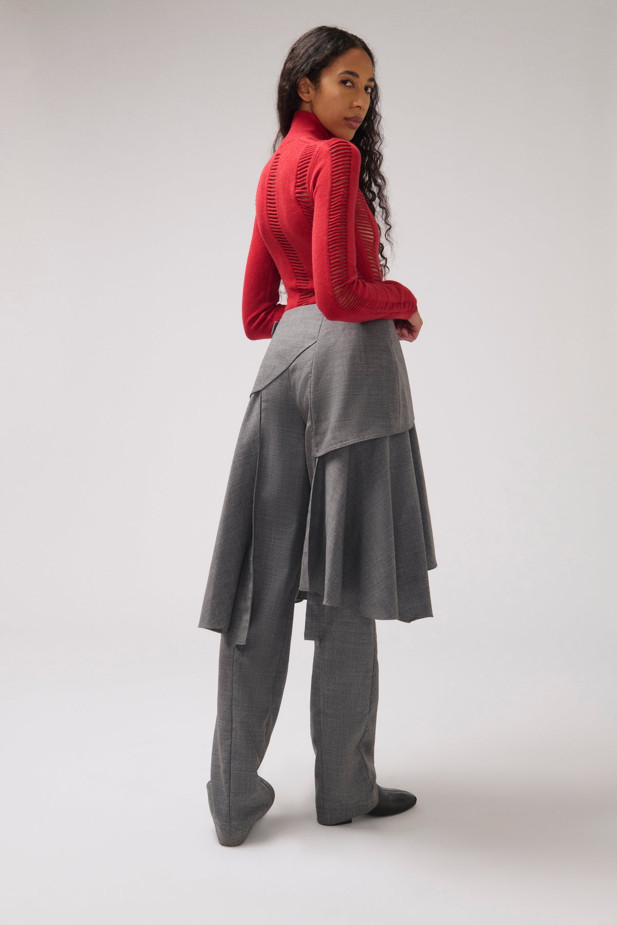 A model is shown from a rear three-quarter angle wearing a bright red ribbed knit mock-neck long-sleeve top and a layered gray draped skirt with wide-leg trousers.