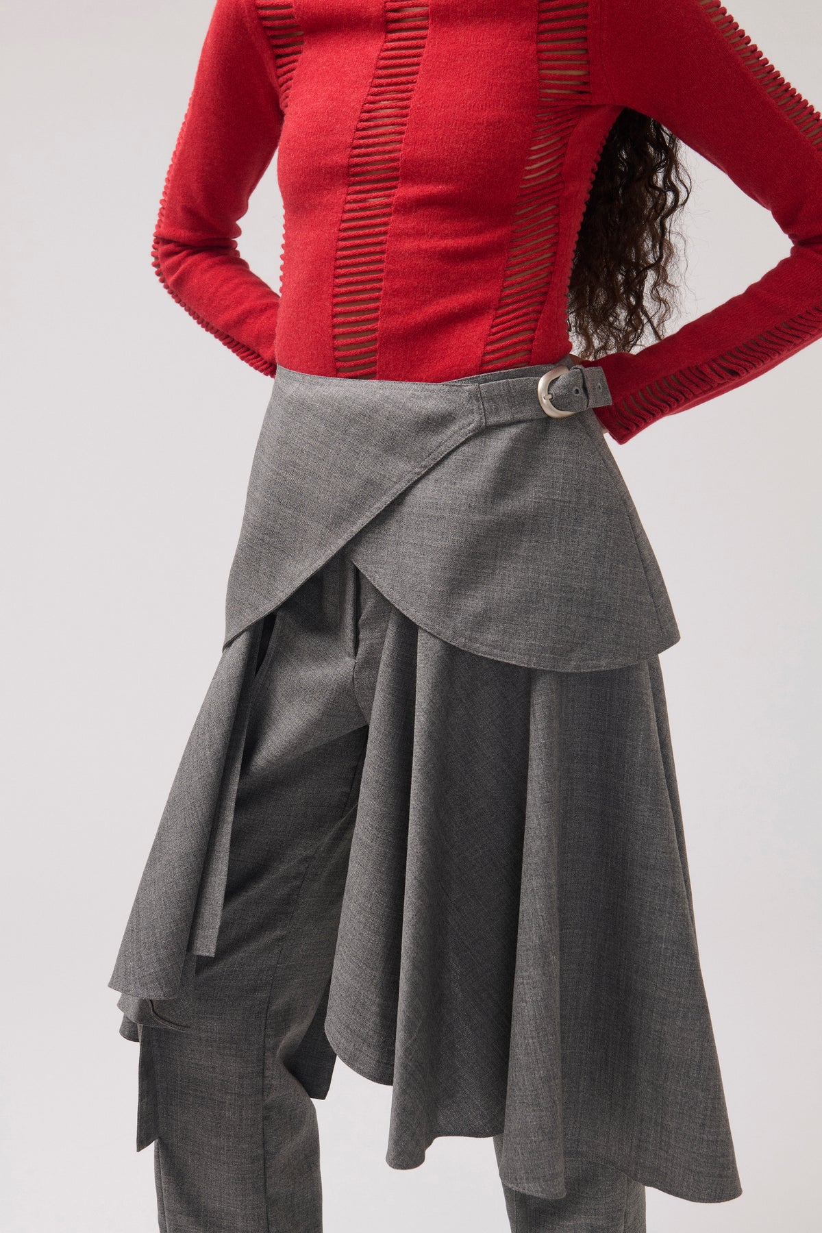 A model wearing a vibrant red ribbed knit top with vertical textured stripes and a gray asymmetrical draped skirt with a single silver button closure at the waist.