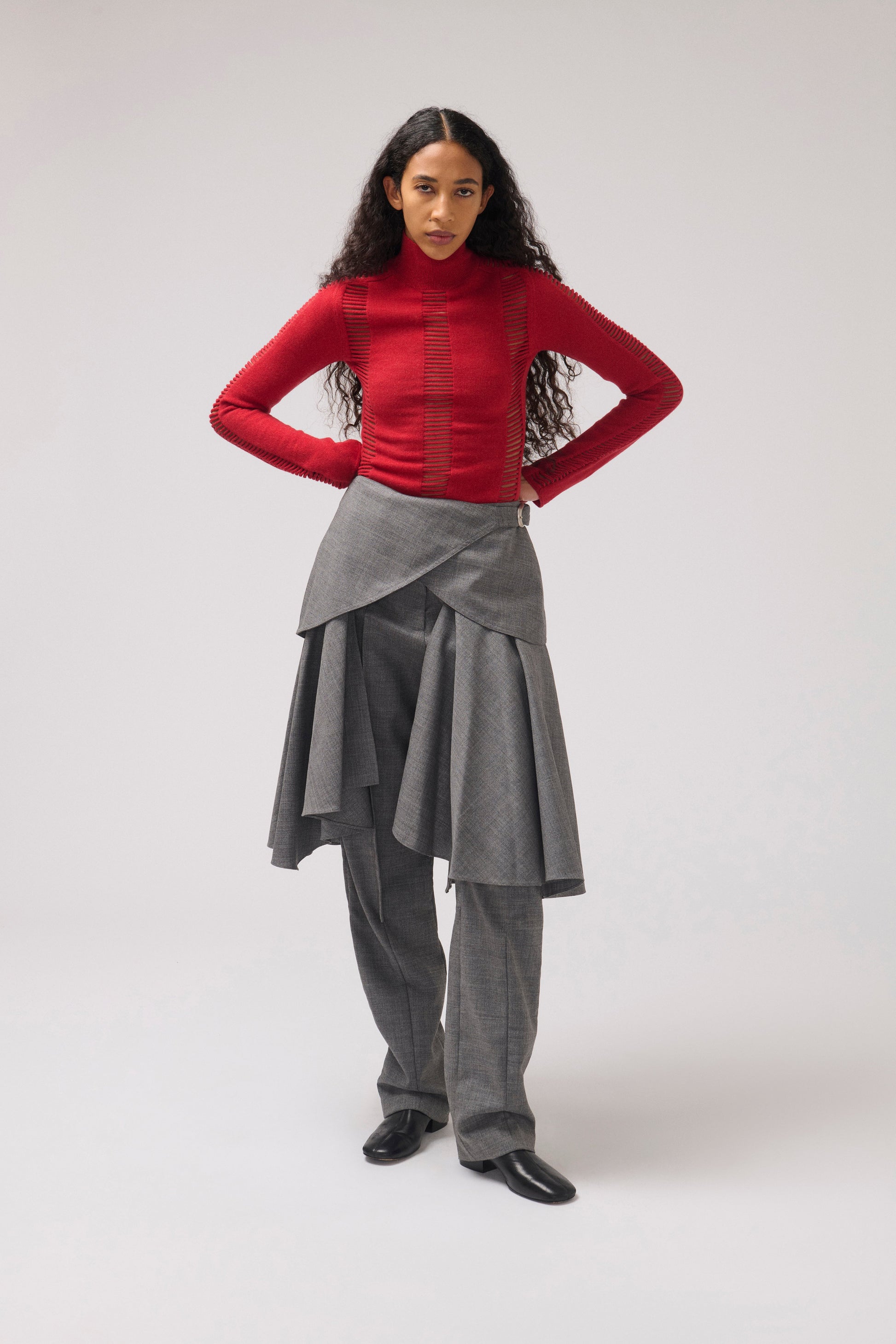 A model stands against a light gray studio background wearing a fitted red long-sleeve mock-neck top, a gray asymmetrical draped trousers, and black shoes.
