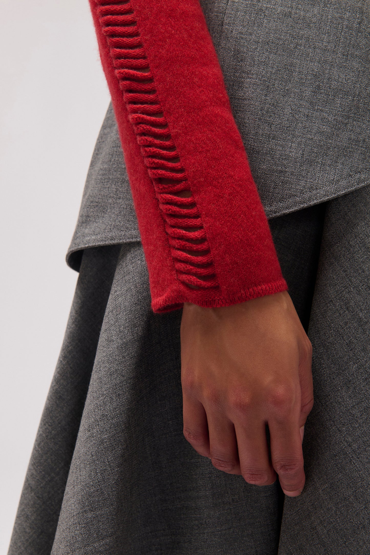 Close-up of a person wearing a textured gray draped garment with a bright red sleeve featuring a vertical ribbed slit detail, with the hand resting near the hem.