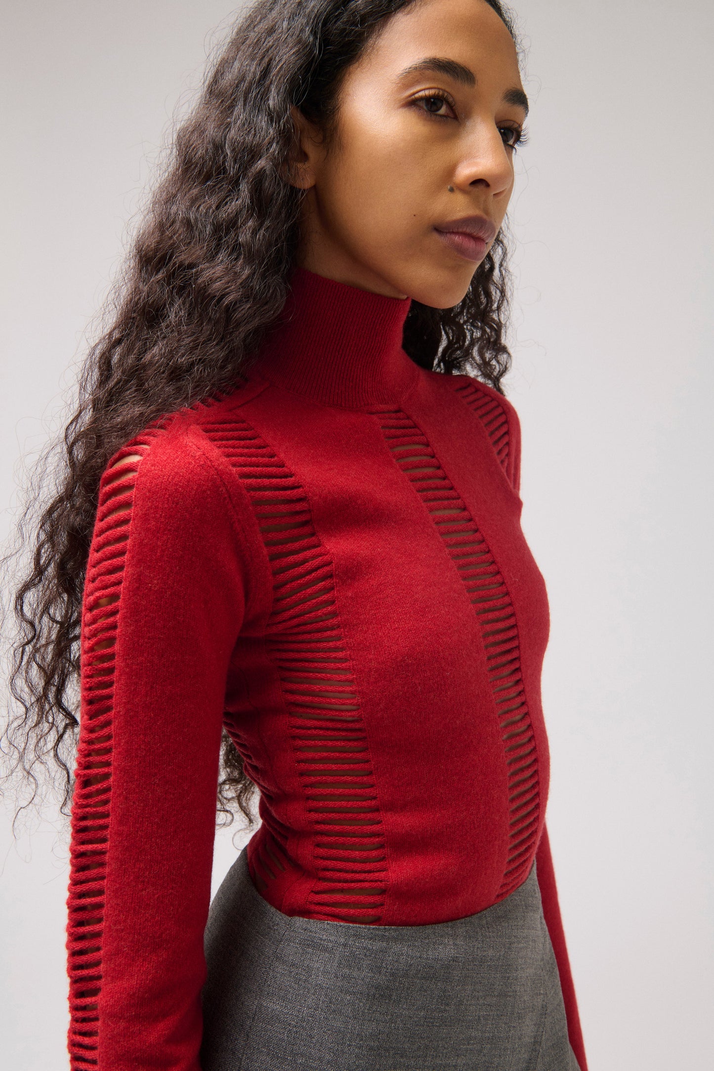A model wearing a red mock-neck knit top with vertical slit details on the chest and sleeves, paired with gray trousers. Photographed against a neutral light gray background.