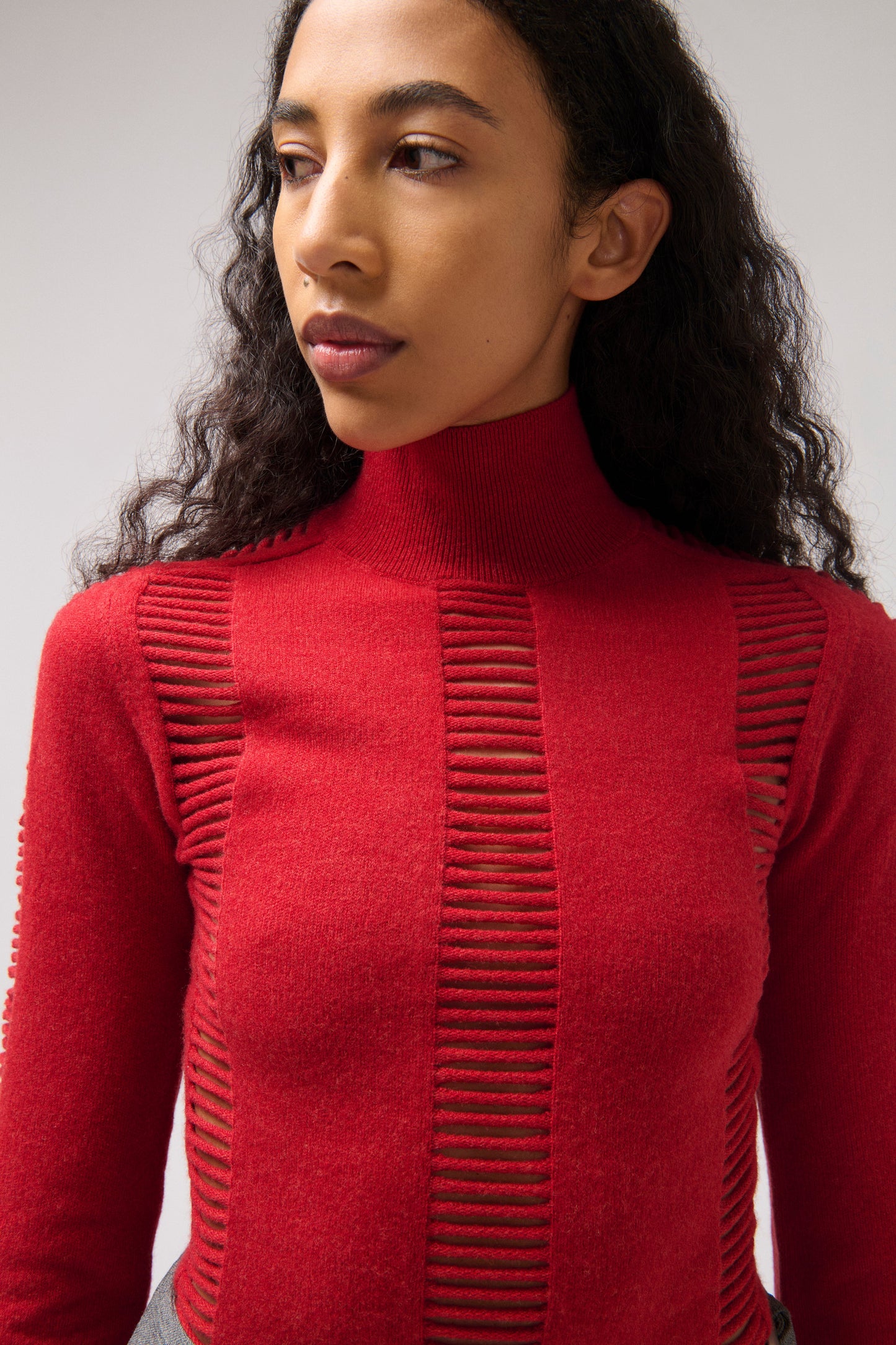 A close-up of a model wearing a vibrant red mock neck knit sweater with vertical ribbed texture details on the chest, set against a plain light gray background.