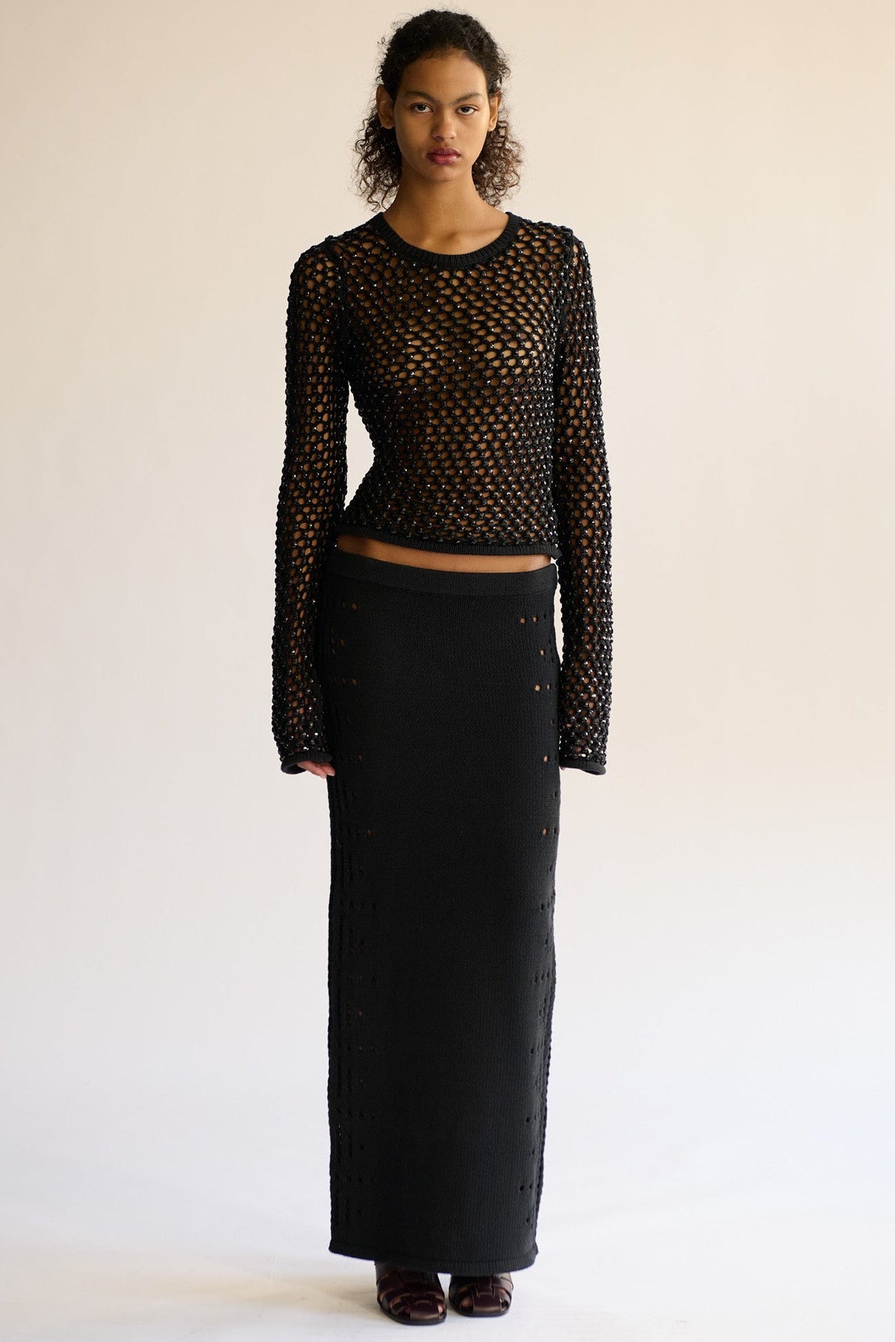 model wearing a black long-sleeve top crystal mesh top and black maxi merino mesh skirt with small cutouts along the side photographed against a white background 