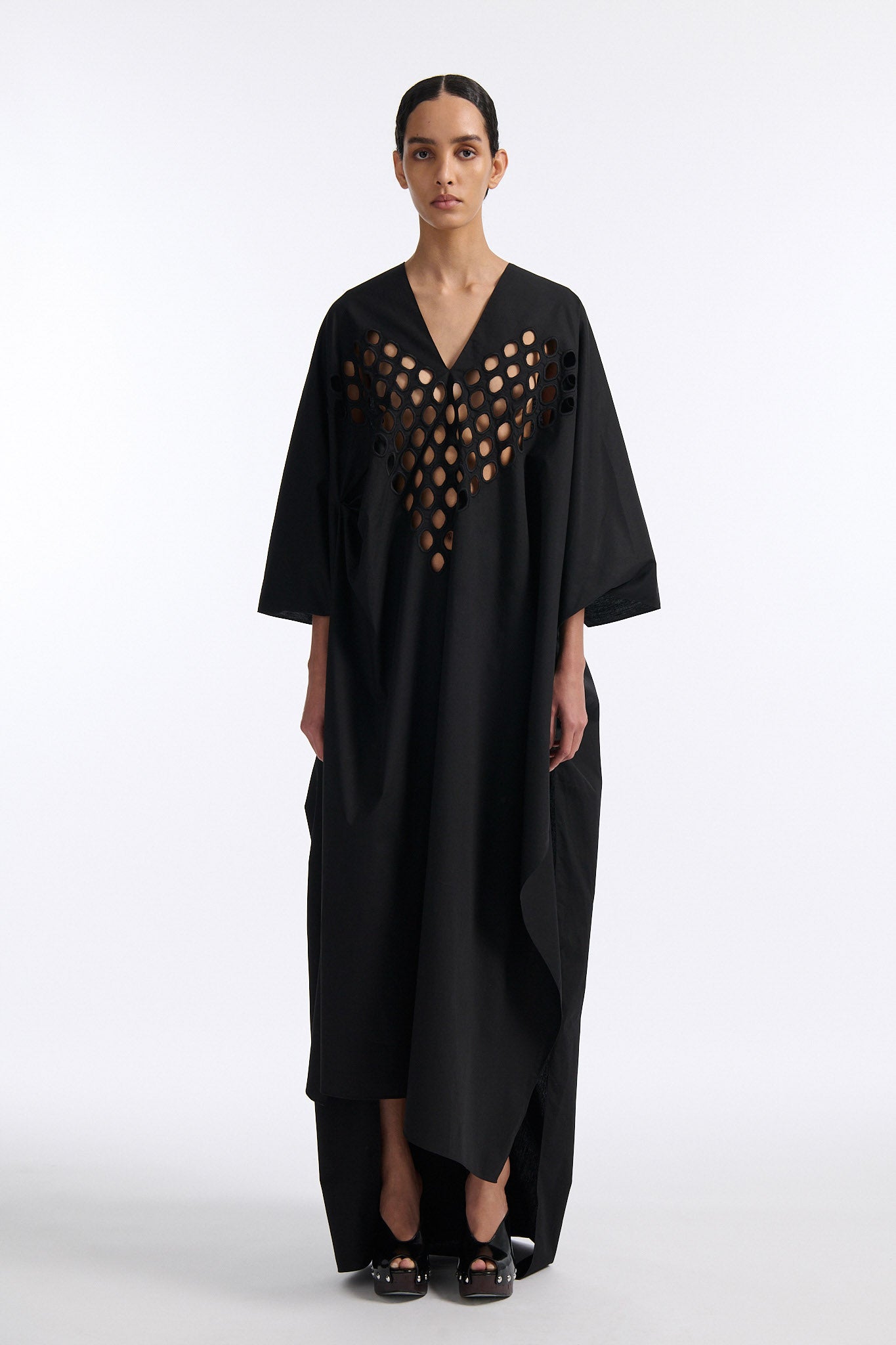 Female model with slicked back black hair in a bun wears black cotton V-neck caftan with rounded diamond cutouts along chest and upper abdomen as well as on the back. The caftan covers both arms and has a shorter front than back with slits along either side. 