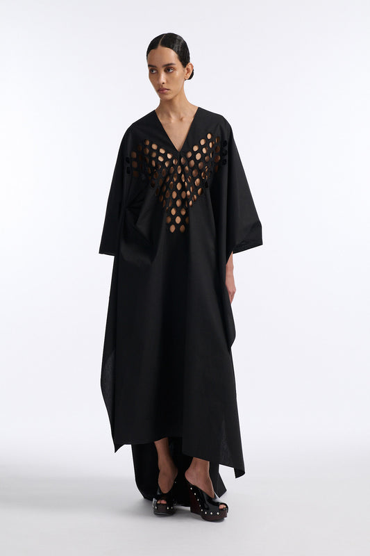 Female model with slicked back black hair in a bun wears black cotton V-neck caftan with rounded diamond cutouts along chest and upper abdomen as well as on the back. The caftan covers both arms and has a shorter front than back with slits along either side. 