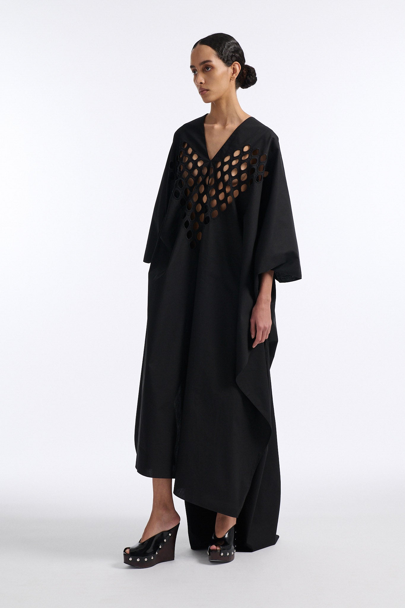 Female model with slicked back black hair in a bun wears black cotton V-neck caftan with rounded diamond cutouts along chest and upper abdomen as well as on the back. The caftan covers both arms and has a shorter front than back with slits along either side. 