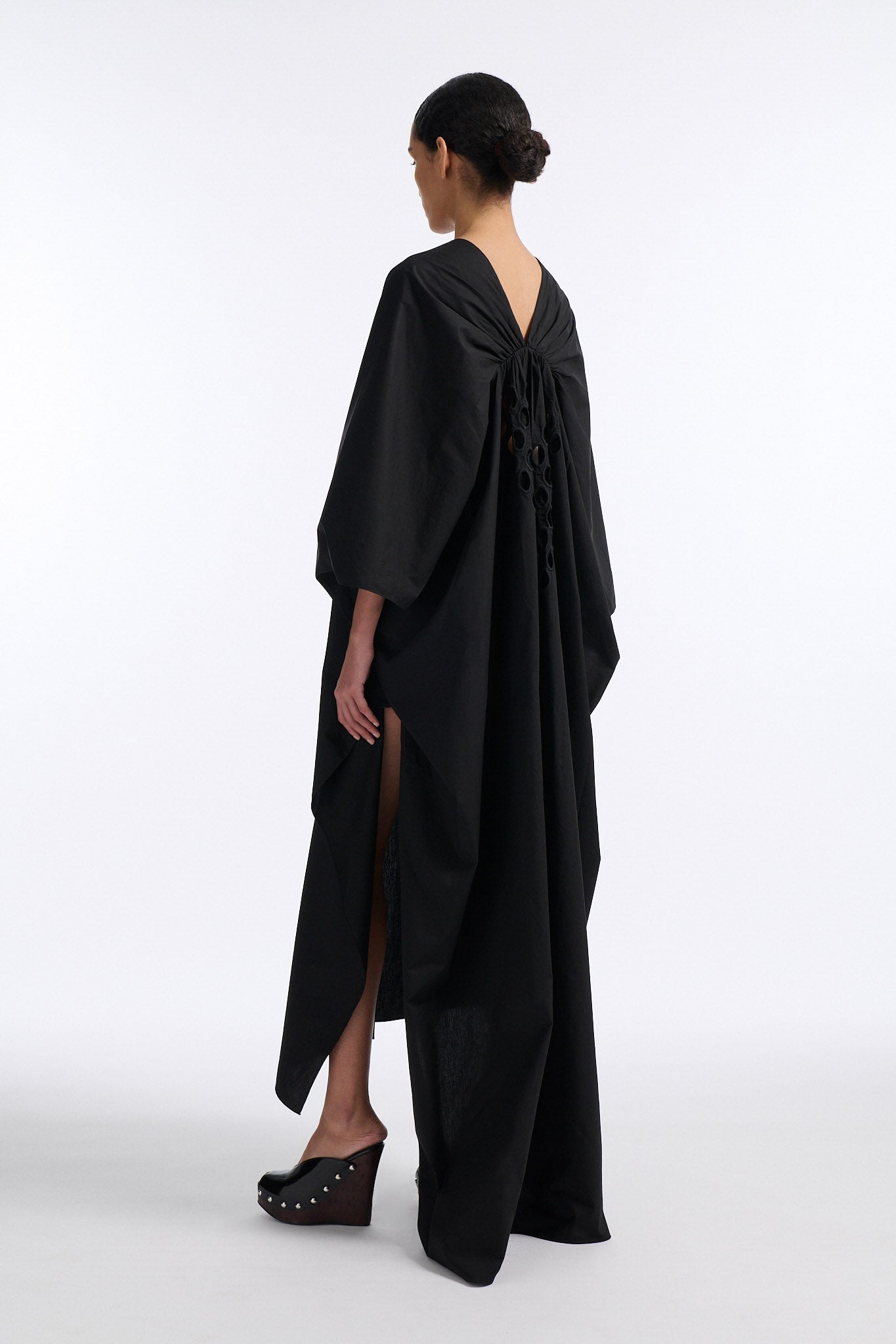 Female model with slicked back black hair in a bun wears black cotton V-neck caftan with rounded diamond cutouts along chest and upper abdomen as well as on the back. The caftan covers both arms and has a shorter front than back with slits along either side. 